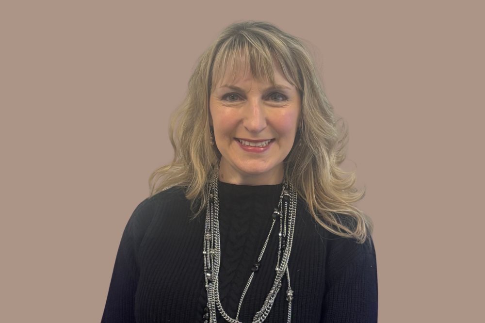 A middle aged woman with long blonde hair and dark eyes is shown smiling at the camera. The woman, Kirsta, is wearing long layered necklaces and a black high neck dress.