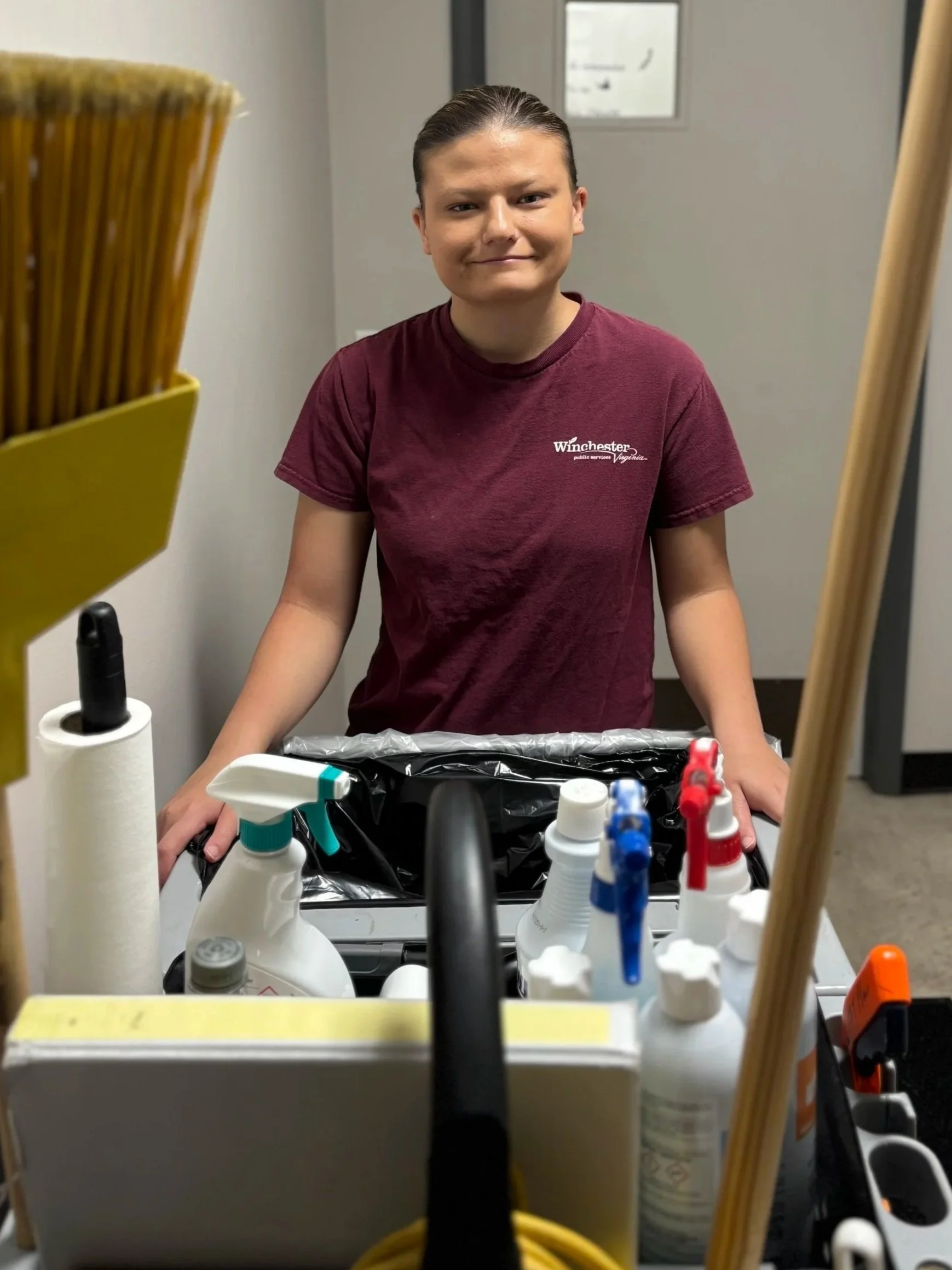 A young woman is shown smiling as she stands behind her custodial cart. The woman has her hair slicked back and is wearing a maroon City of Winchester t-shirt.