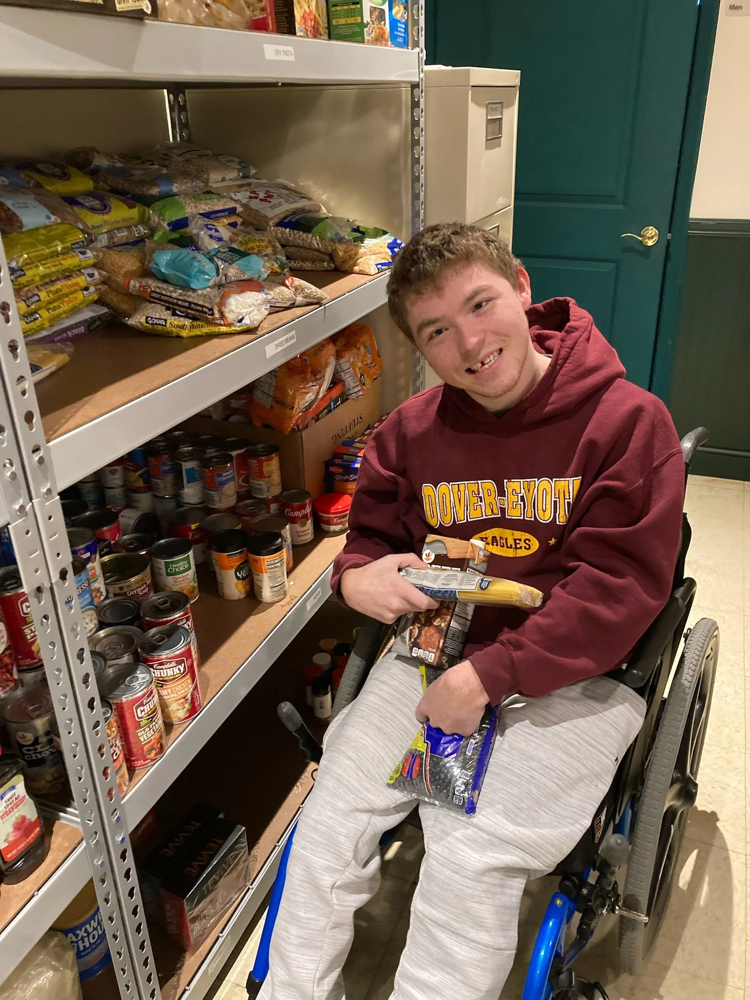 A man in a maroon sweatshirt sits in his wheelchair in front of a row of shelving that holds nonperishable food items. He has more food in his arms and is smiling at the camera.