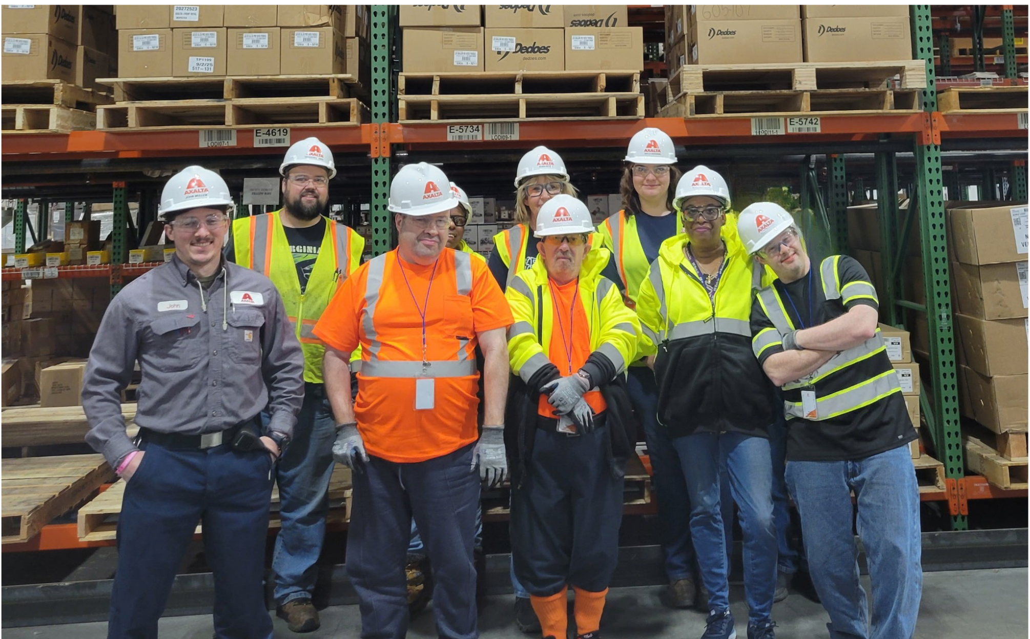 A group of NW Works team members and a job coach stand with employees at Axalta. All people in the picture are wearing hard hats and some form of a protective or approved vest.