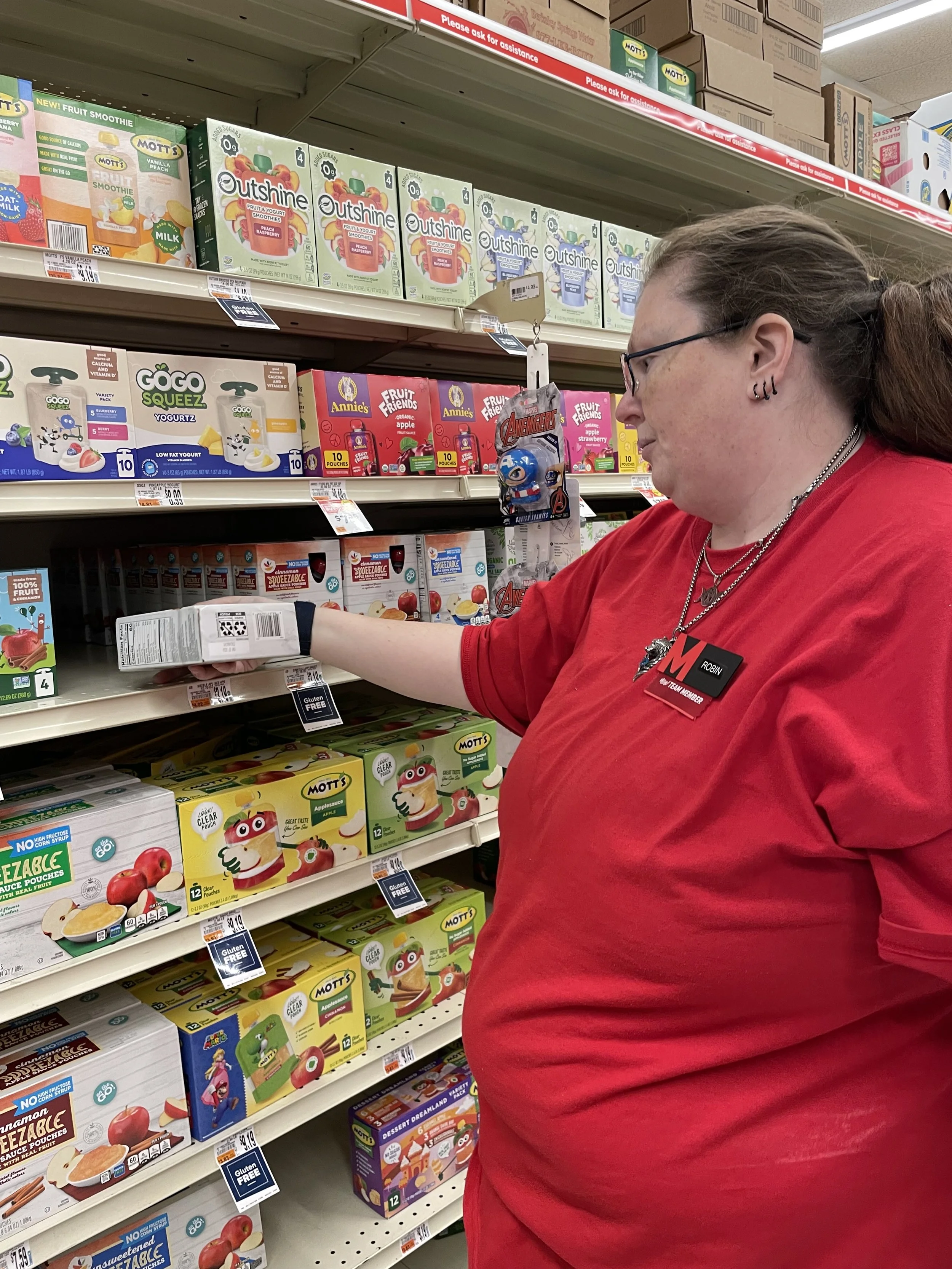 A middle aged woman is shown checking the dates on a package of apple sauce in a grocery store aisle. The woman is wearing a red t-shirt and her Martin's name badge.