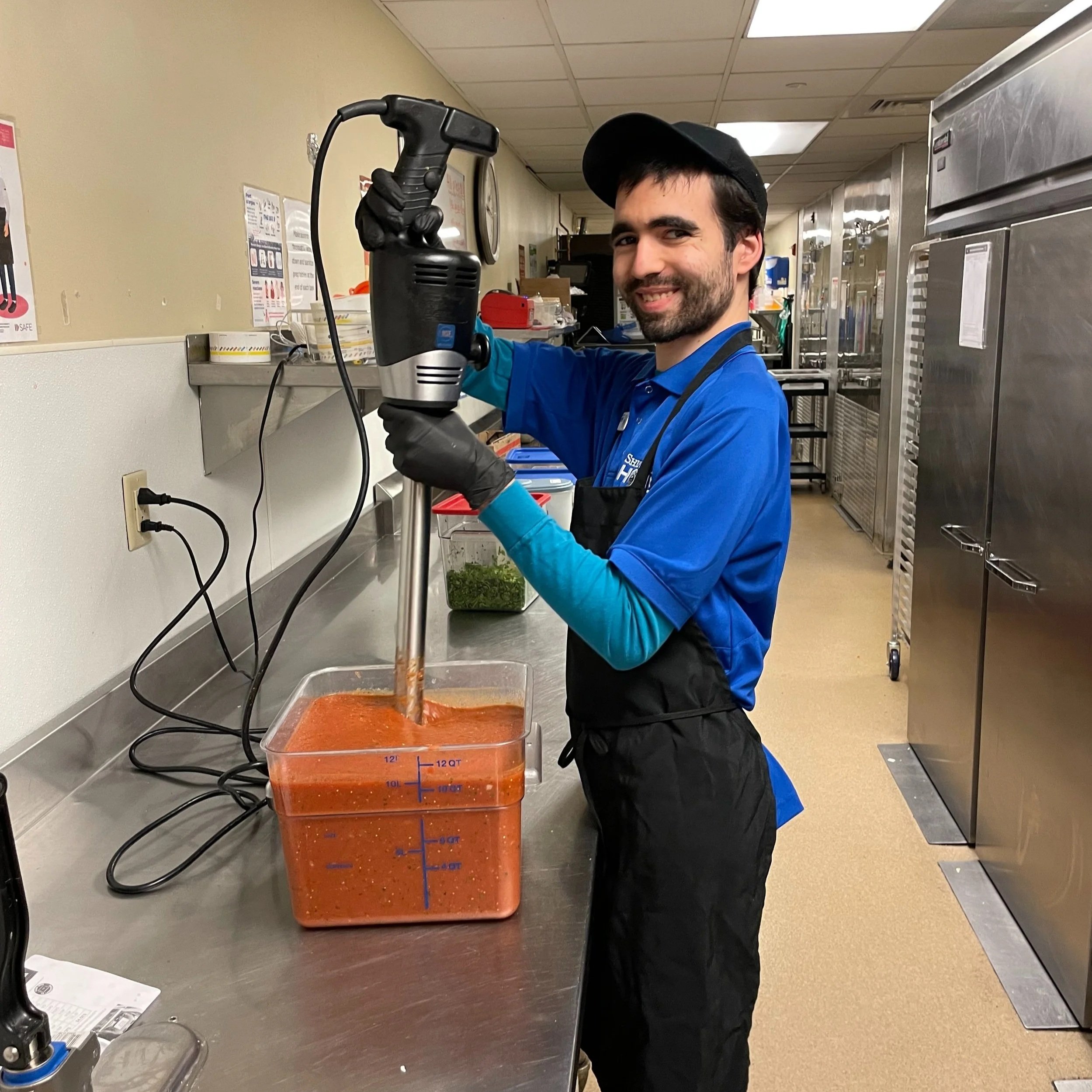 A young man is shown holding an industrial mixer with both hands as he smiles at the camera. The mixer is inserted into a tub of red sauce that is likely salsa. Behind the man an industrial kitchen can be seen.