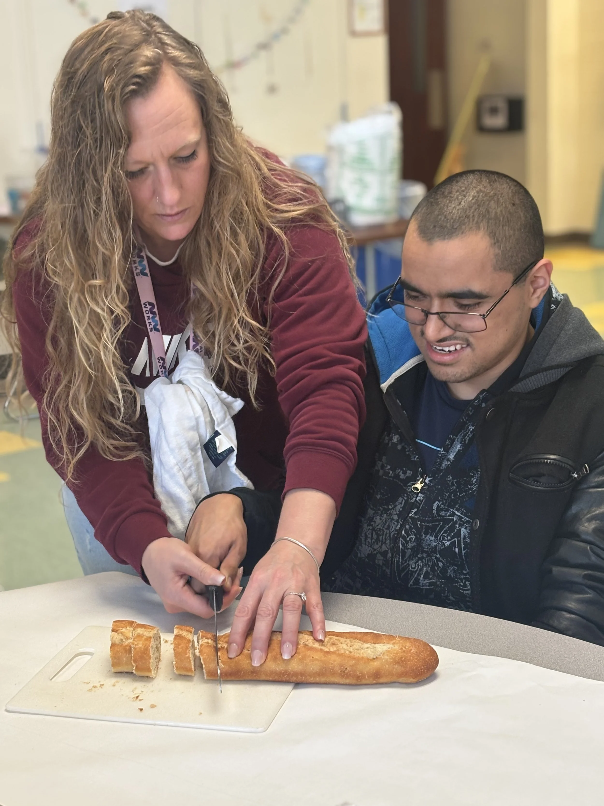 A middle aged woman helps a young man slice a loaf of bread. They are both concentrating on the knife and the bread in front of them.
