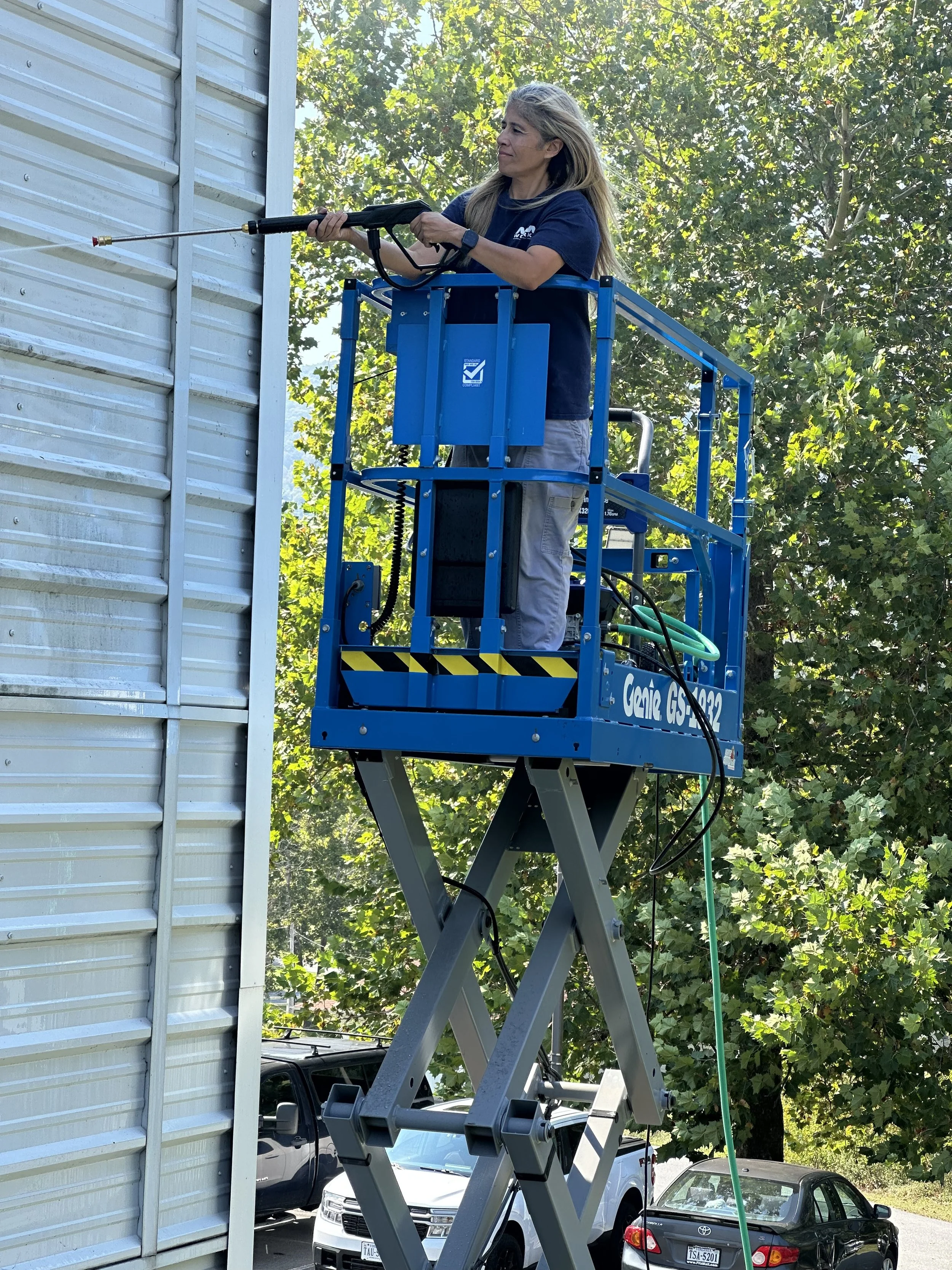  A middle aged woman is shown using a power washer as she stands on a blue scissor lift that has lifted her to clean the outside of a metal building. There are cars below her and she is set up in a parking lot. 