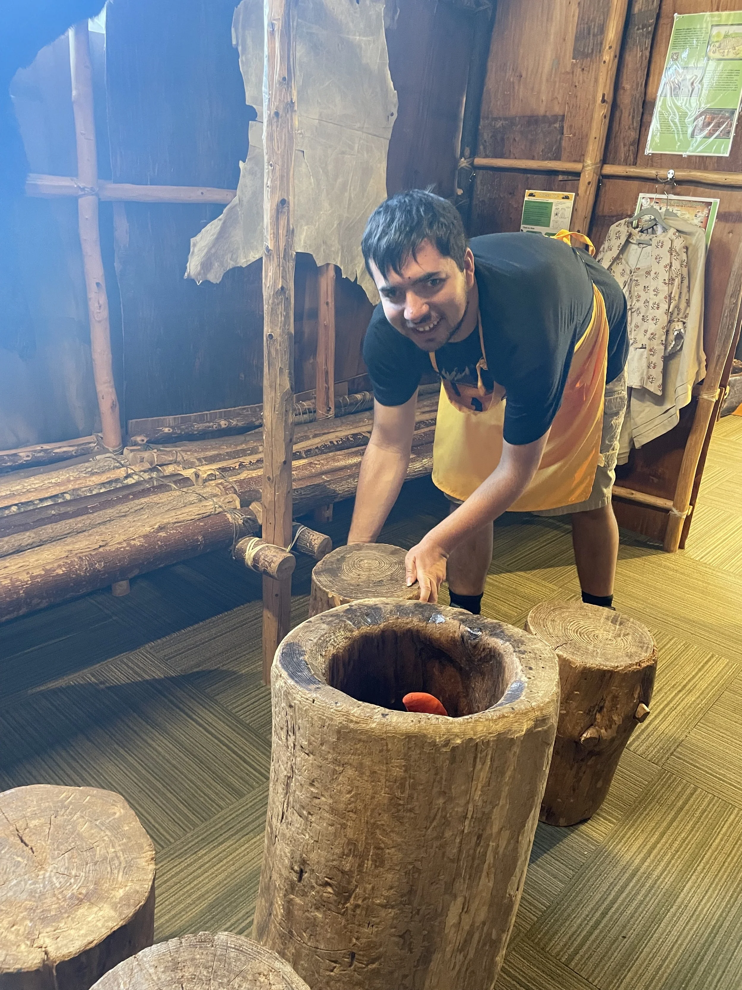 A young man, Peter, is shown moving wooden logs around a room that is designed to look like a colonial home with a wooden bed or bench and older style clothing hung on hangers for dress-up opportunities.