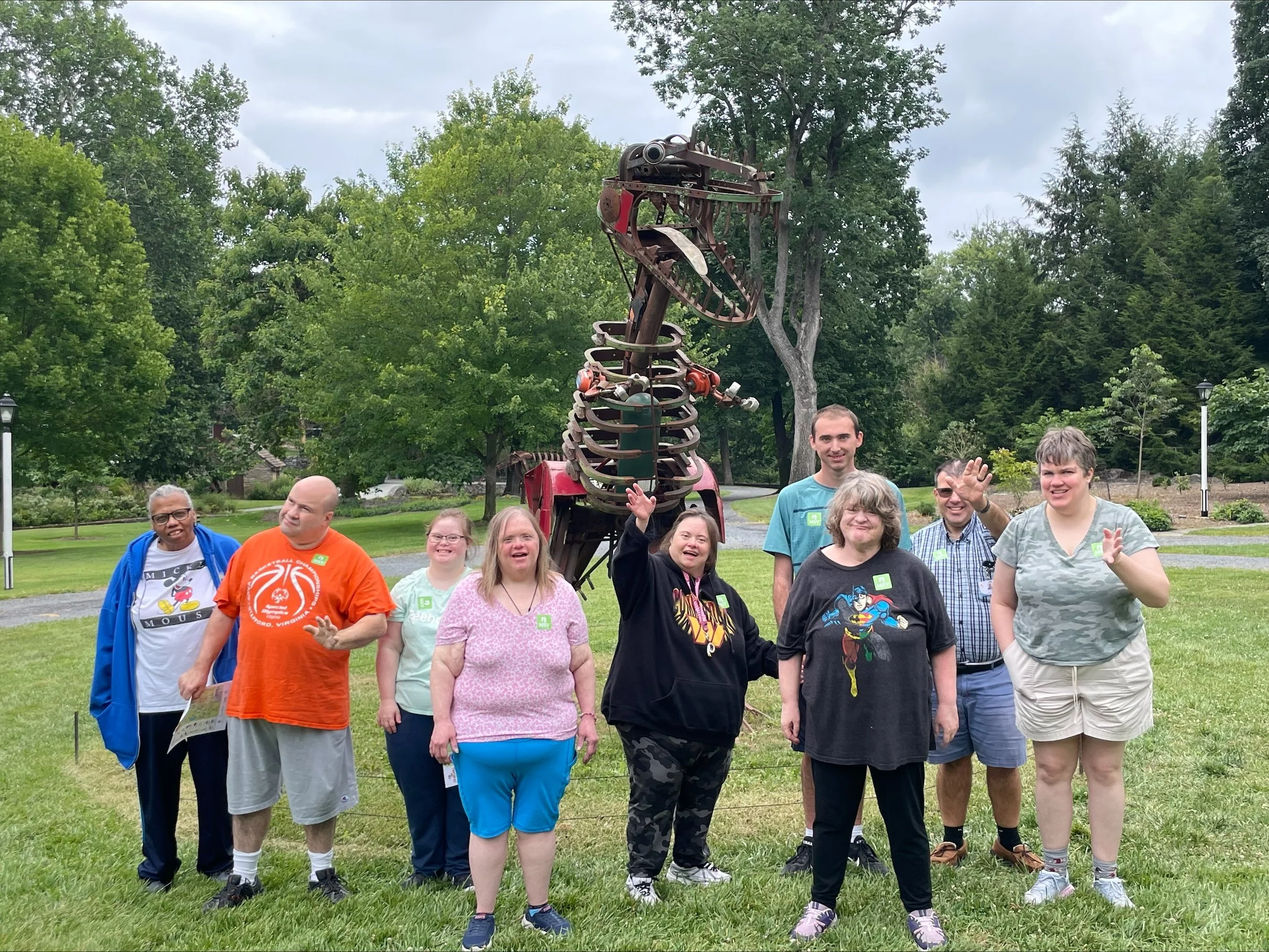 A large group of NW Works team members stand outside in front of a scrap metal dinosaur. Many members of the group are waving or smiling.