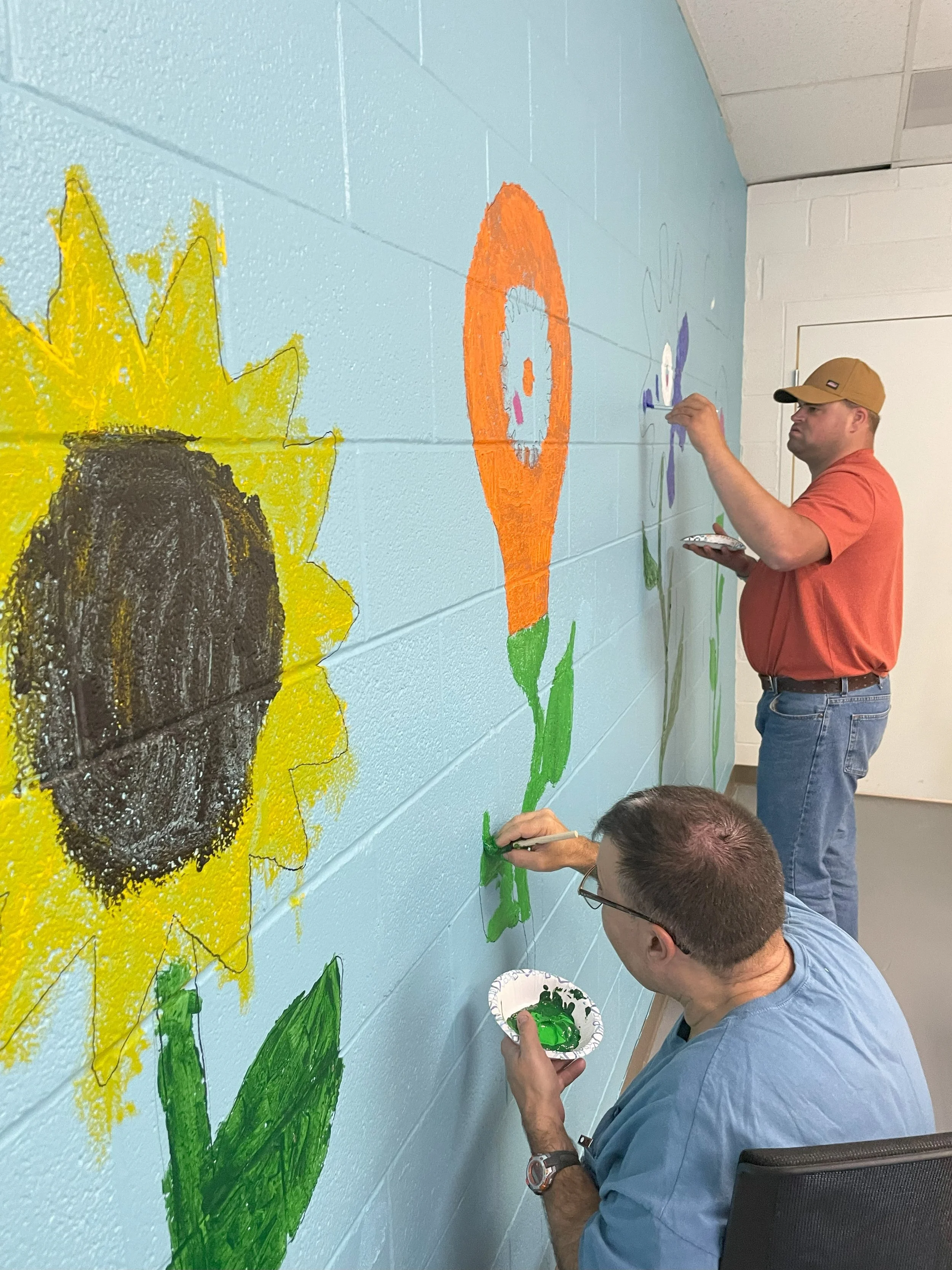 Two men are shown painting flowers with gears for their blooms. The painting is taking place in NW Works' Digital Literacy  Lab.