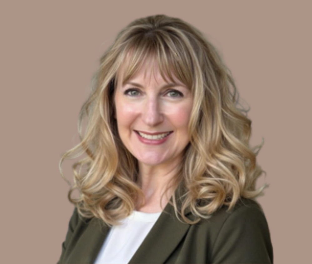 A middle aged woman, Kirsta, is shown. Kirsta has blonde curly hair with brown highlights. She has light colored eyes, and is wearing a white blouse with an olive green blazer over it. The background of her headshot is tan.