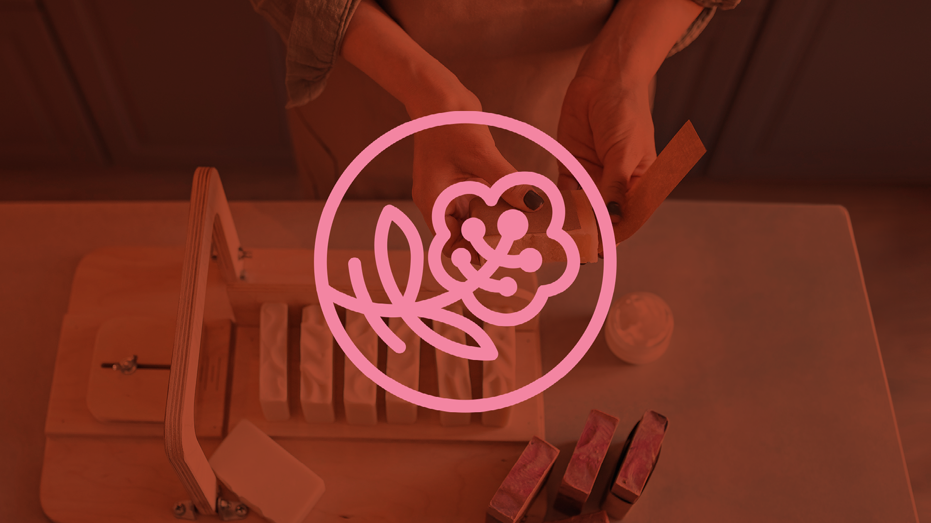 Modern pink line‑art flower logo centered over a rust‑tinted photo of a woman labeling handcrafted soap.