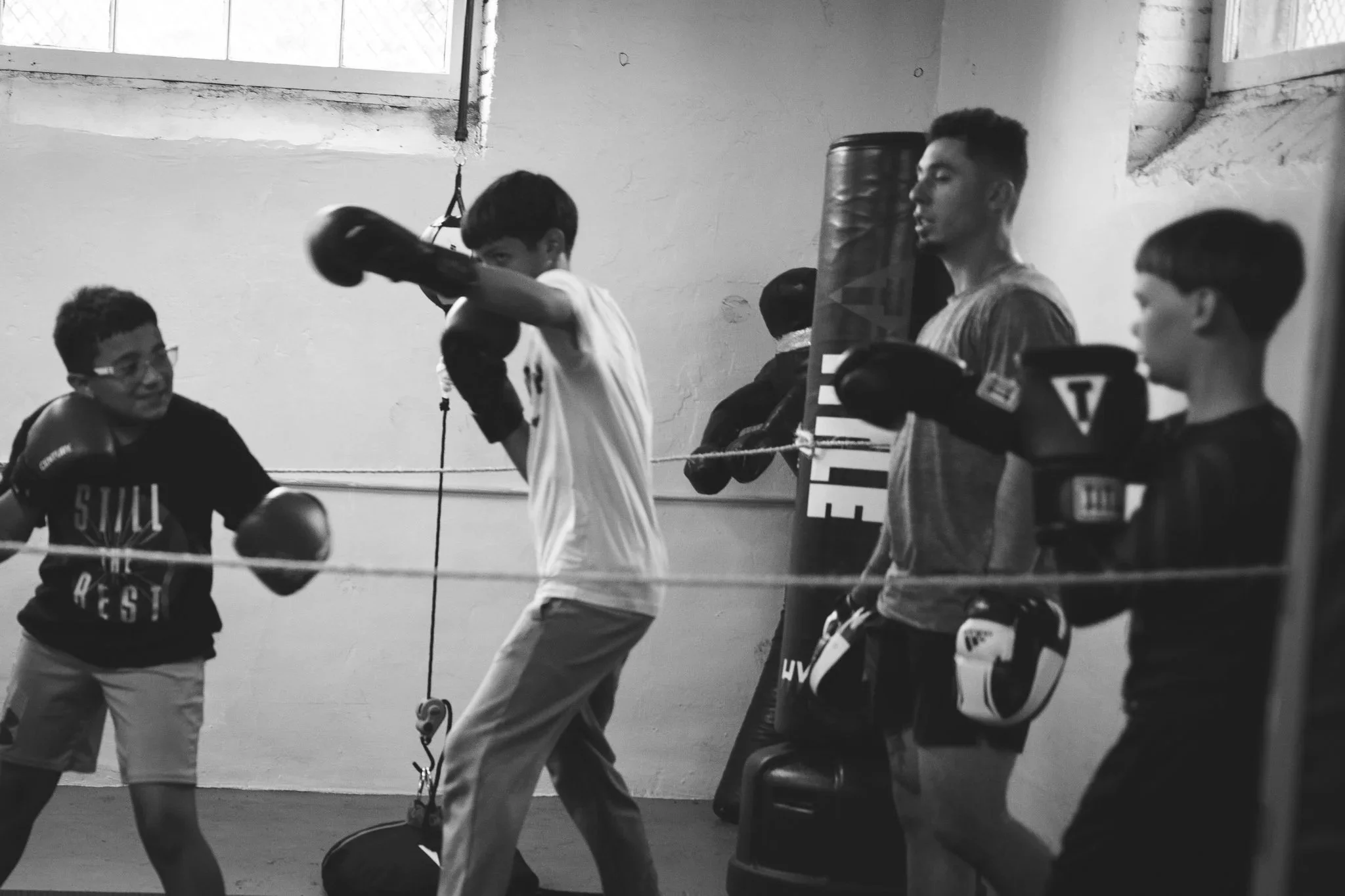 Boxing practice session with five young boys training with punching bags and gloves in a gym with a plain wall and window