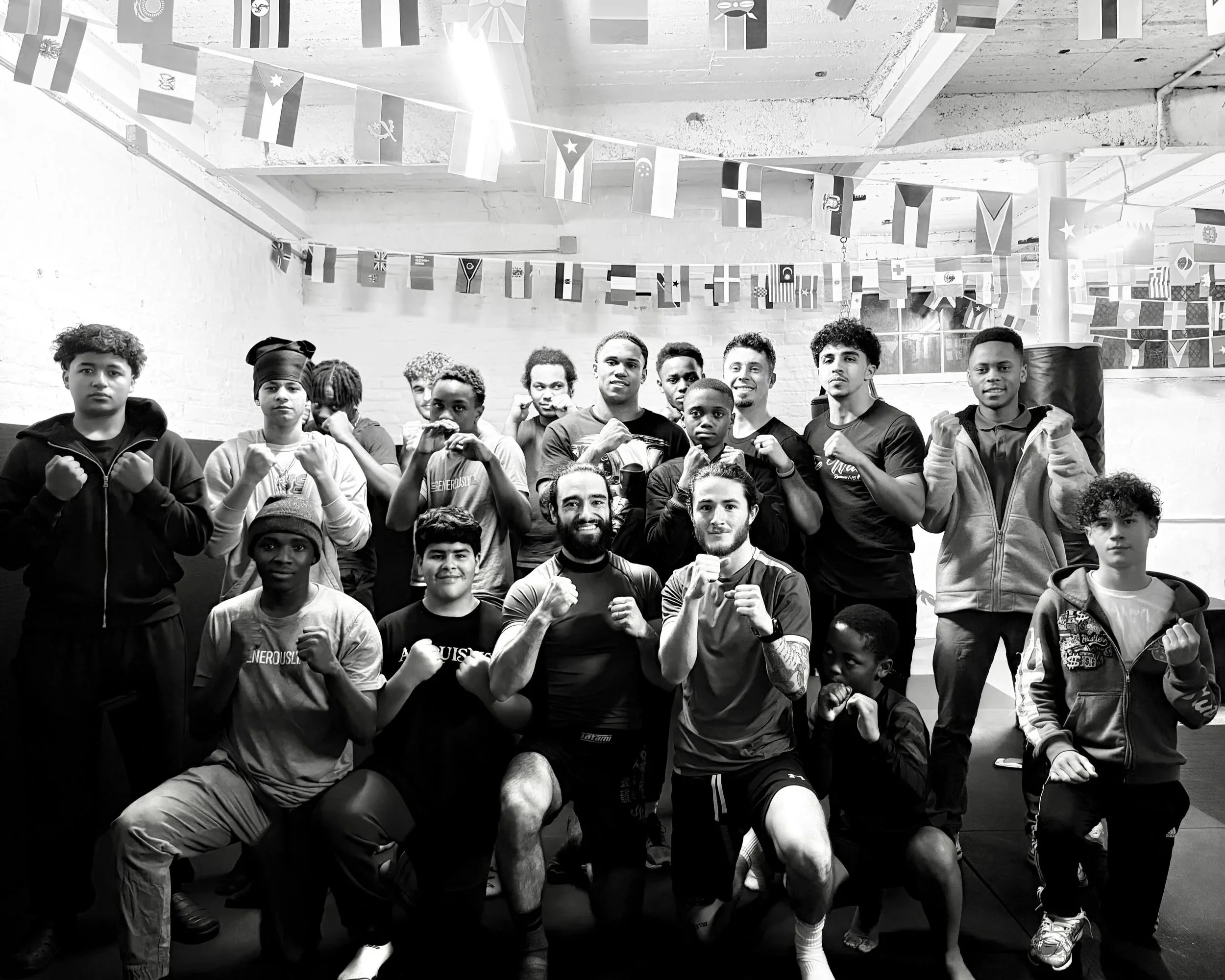 Group of young people and two adult men in athletic stances with fists raised, inside a boxing gym with flags hanging from the ceiling.