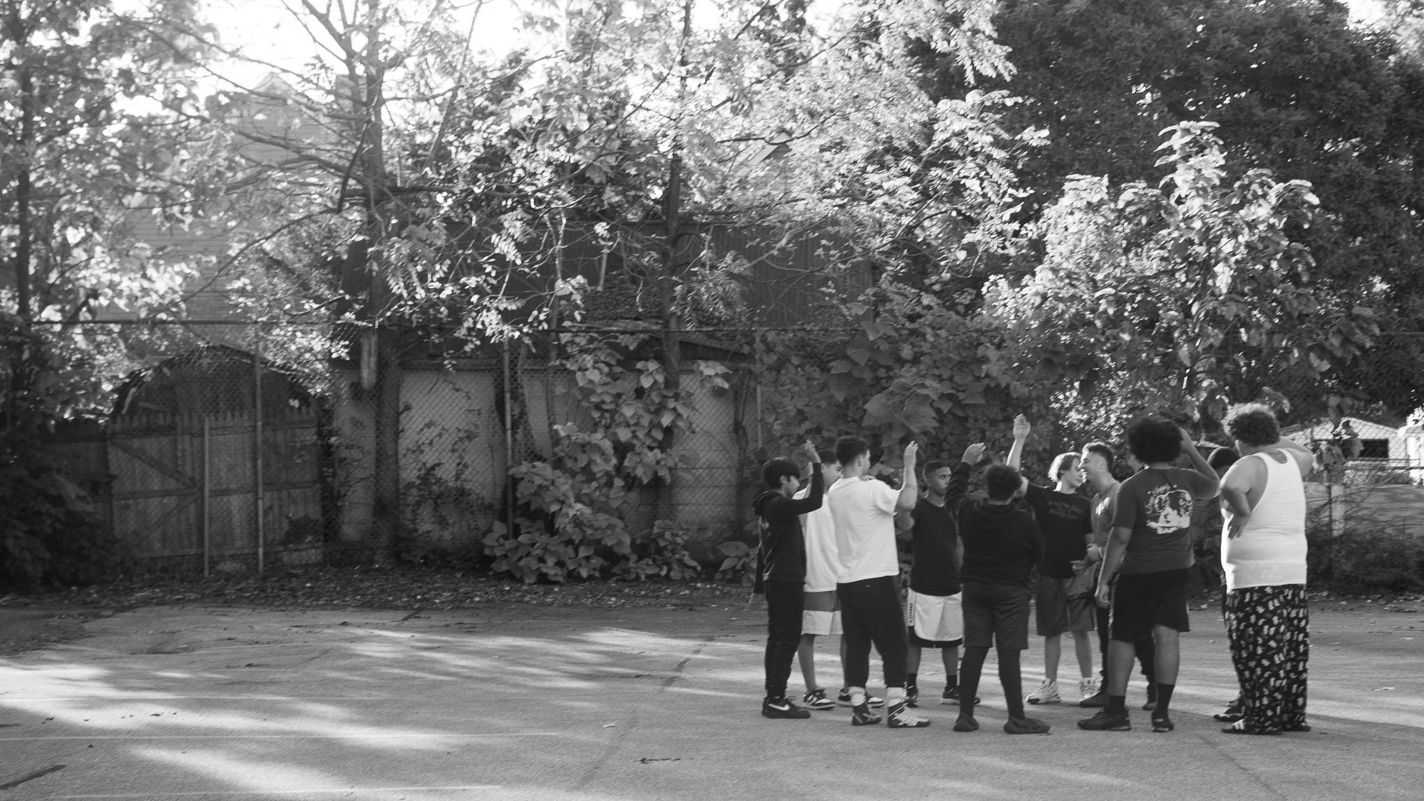 A group of children and teenagers gather on a street, appearing to play or chat, with trees and a fence in the background.