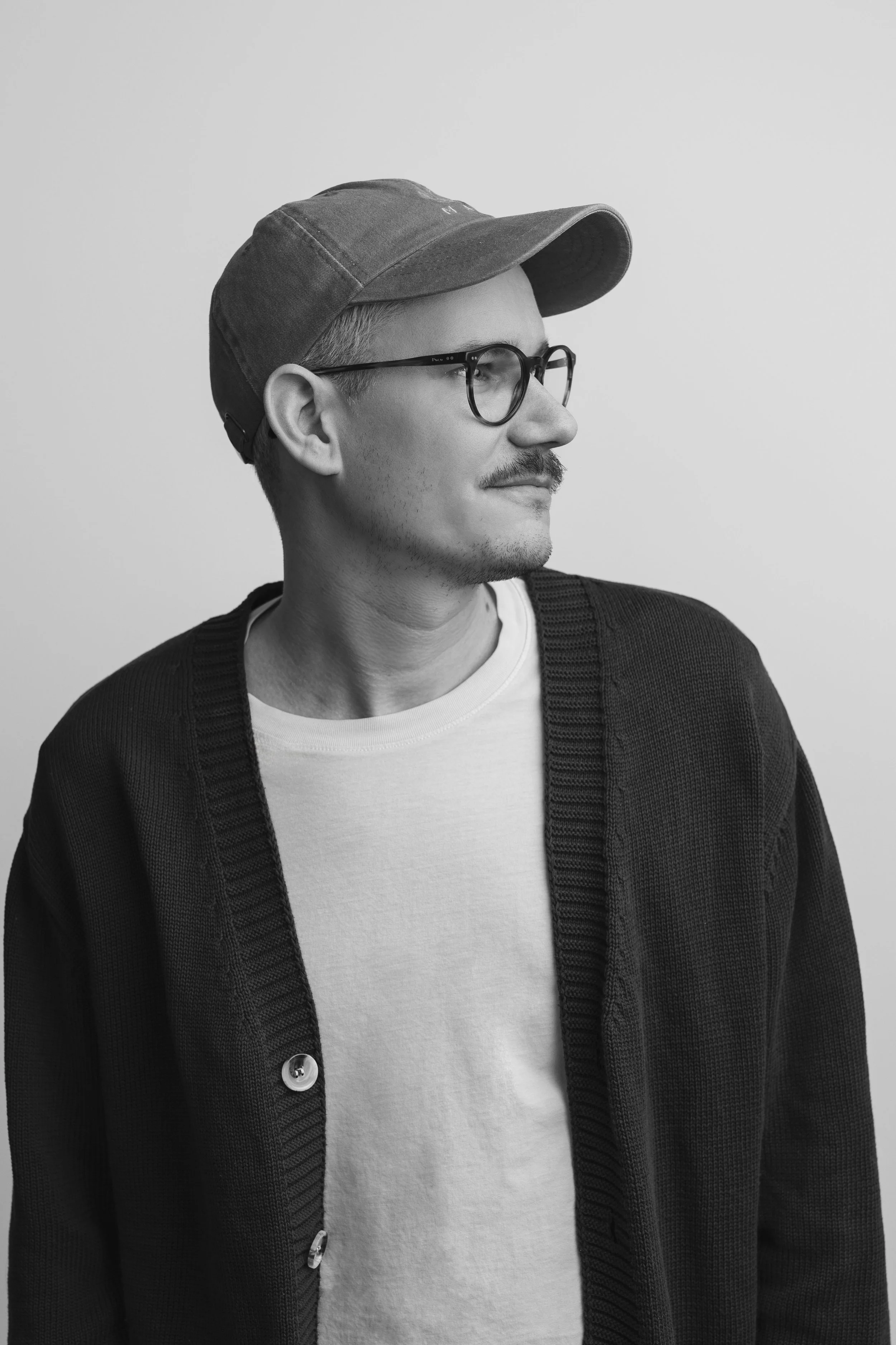 Man wearing glasses and a cap, looking to the side, dressed in a cardigan and t-shirt, black and white photo.