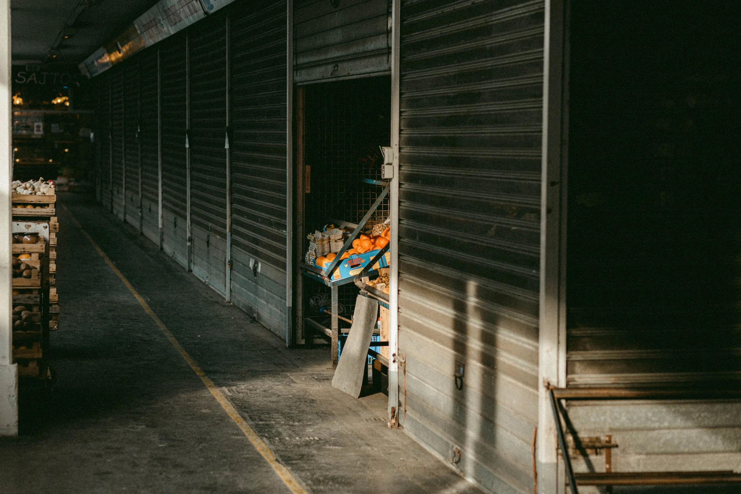 Dimly lit indoor market with closed metal doors and a small open stall displaying oranges and other produce in crates.