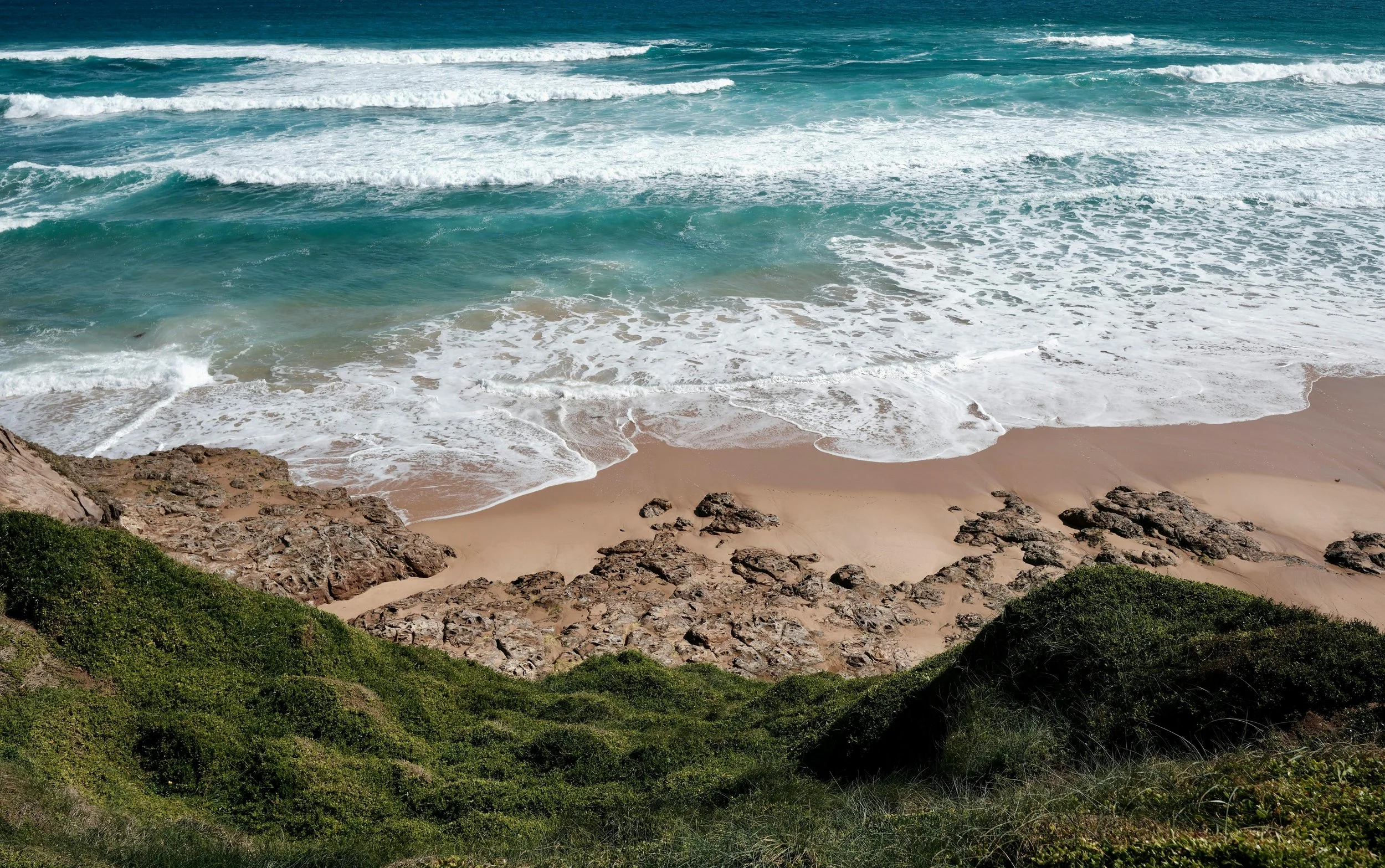 A view from above of turquoise waves meeting a sandy beach, with rocky edges and green grass in the foreground. The ocean carries a sense of renewal and nourishment.