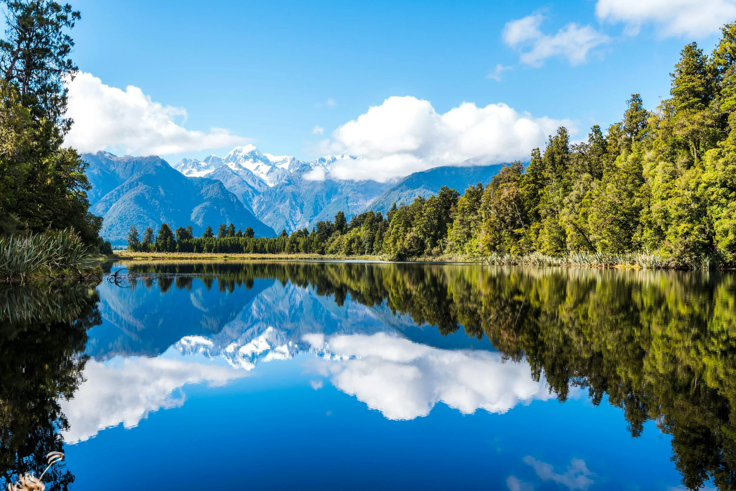 A breathtaking view of snow-capped mountains reflected in a crystal-clear lake, surrounded by lush green forest, showcasing the beauty of untouched nature.