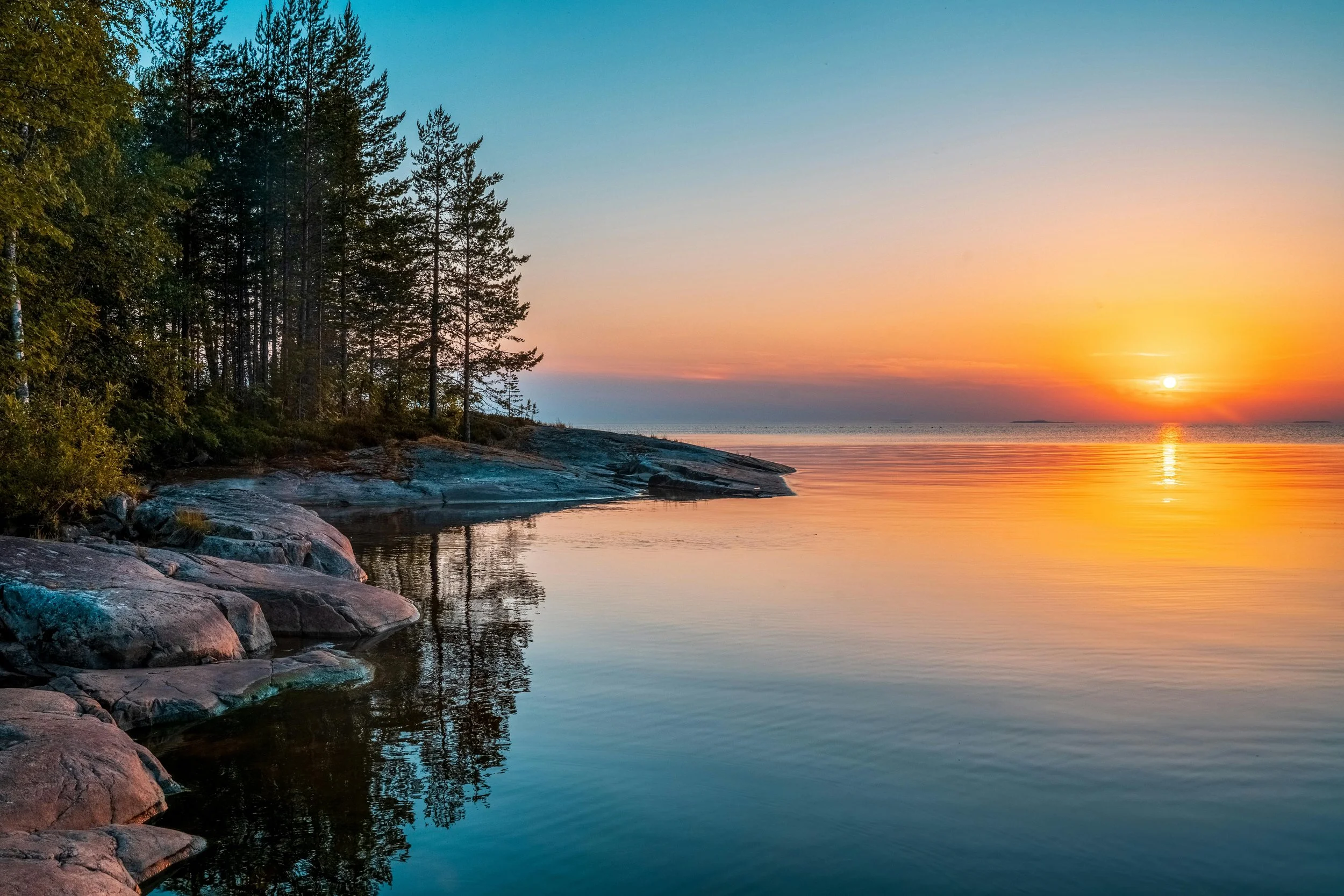 Tranquil sunset over a calm lake with rocky shoreline and pine trees, symbolizing peace and connection with nature.