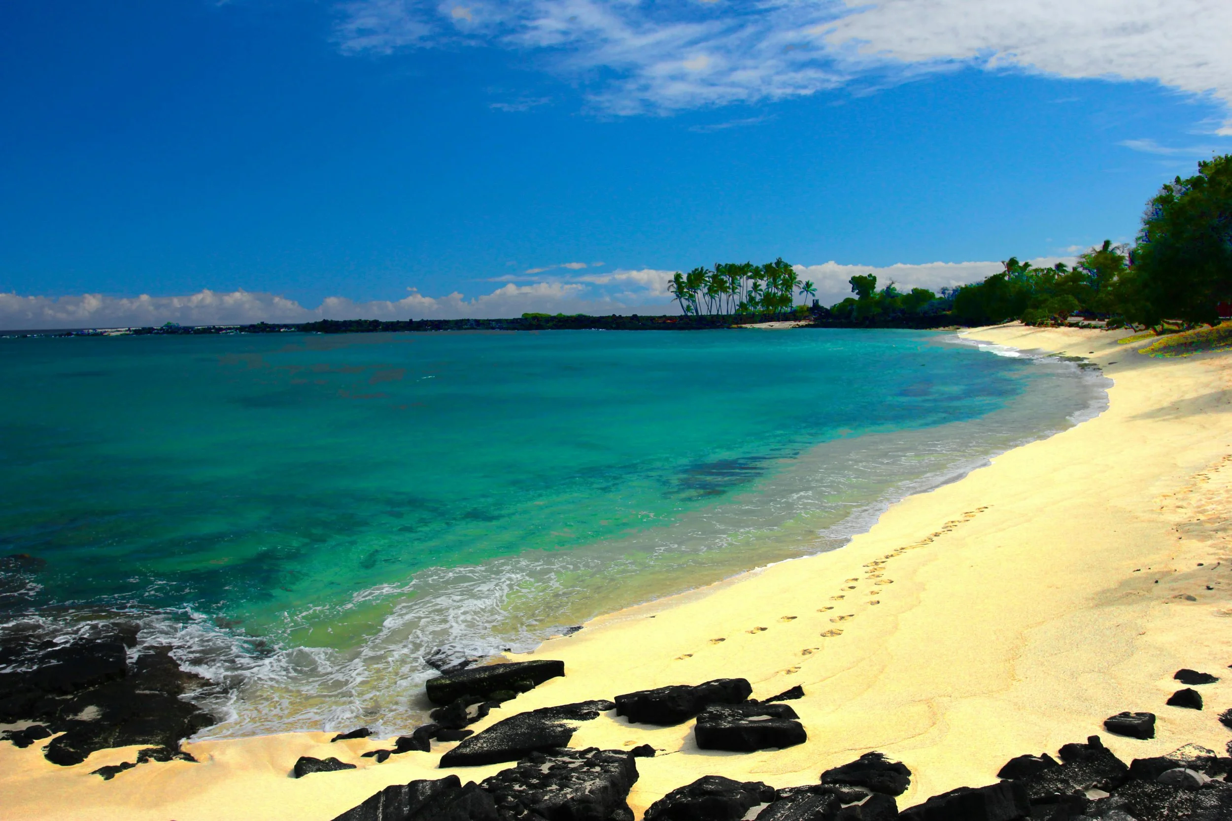 A golden sandy beach with turquoise waves gently washing ashore, leaving footprints in the sand. Dark volcanic rocks frame the shoreline, while palm trees cluster in the distance under a bright blue sky with scattered clouds.
