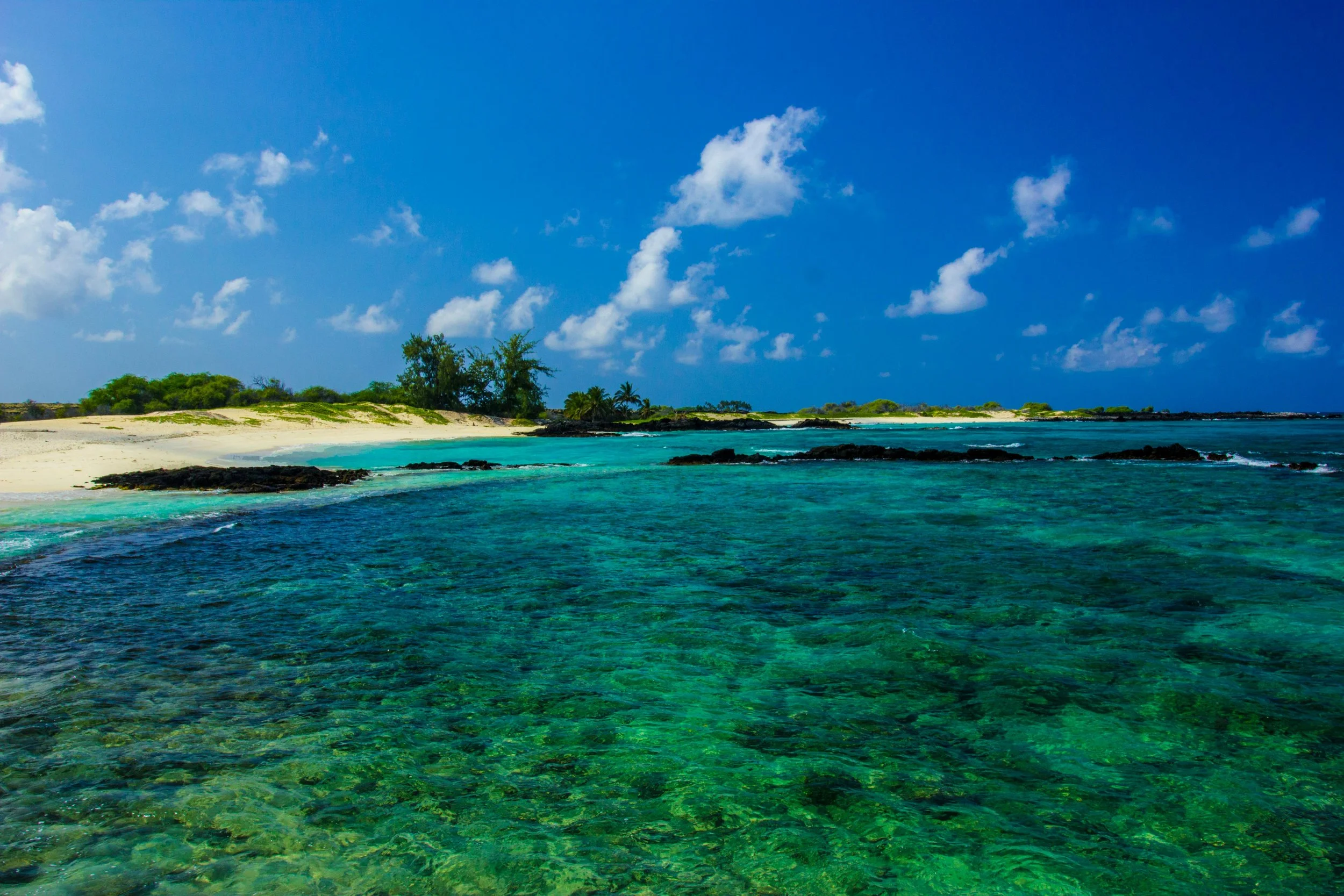A tropical beach with crystal-clear turquoise waters, white sandy shore, and lush greenery under a bright blue sky with scattered clouds.