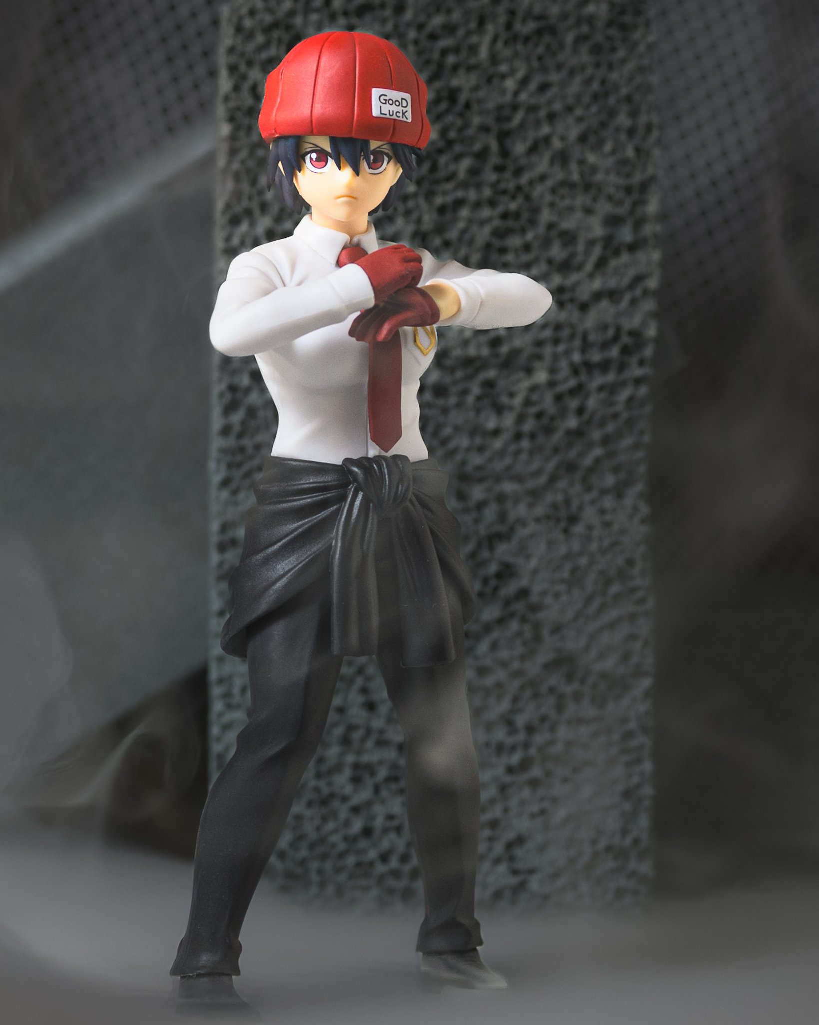 Figure of Fuuko Izumo from Undead Unluck standing in a determined pose with crossed red-gloved hands. She wears a red "Good Luck" beanie, white shirt, and red tie, framed by drifting fog in the foreground and textured stone pillars in the background.