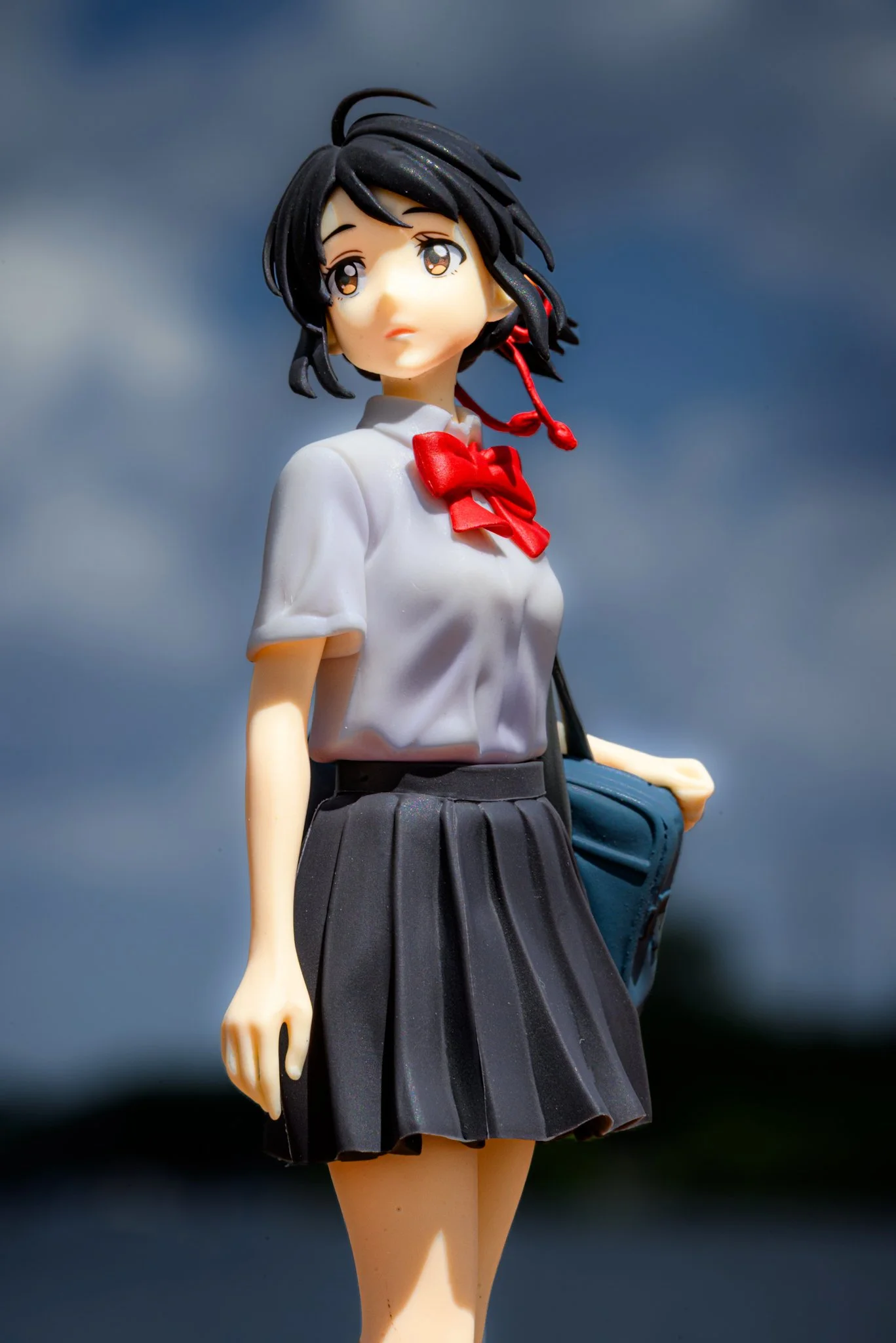 Anime-style schoolgirl figure photographed outdoors against a soft blue sky, wearing a white blouse with a red bow and a dark pleated skirt, holding a bag over her shoulder and looking slightly upward with sunlight casting gentle shadows across her f