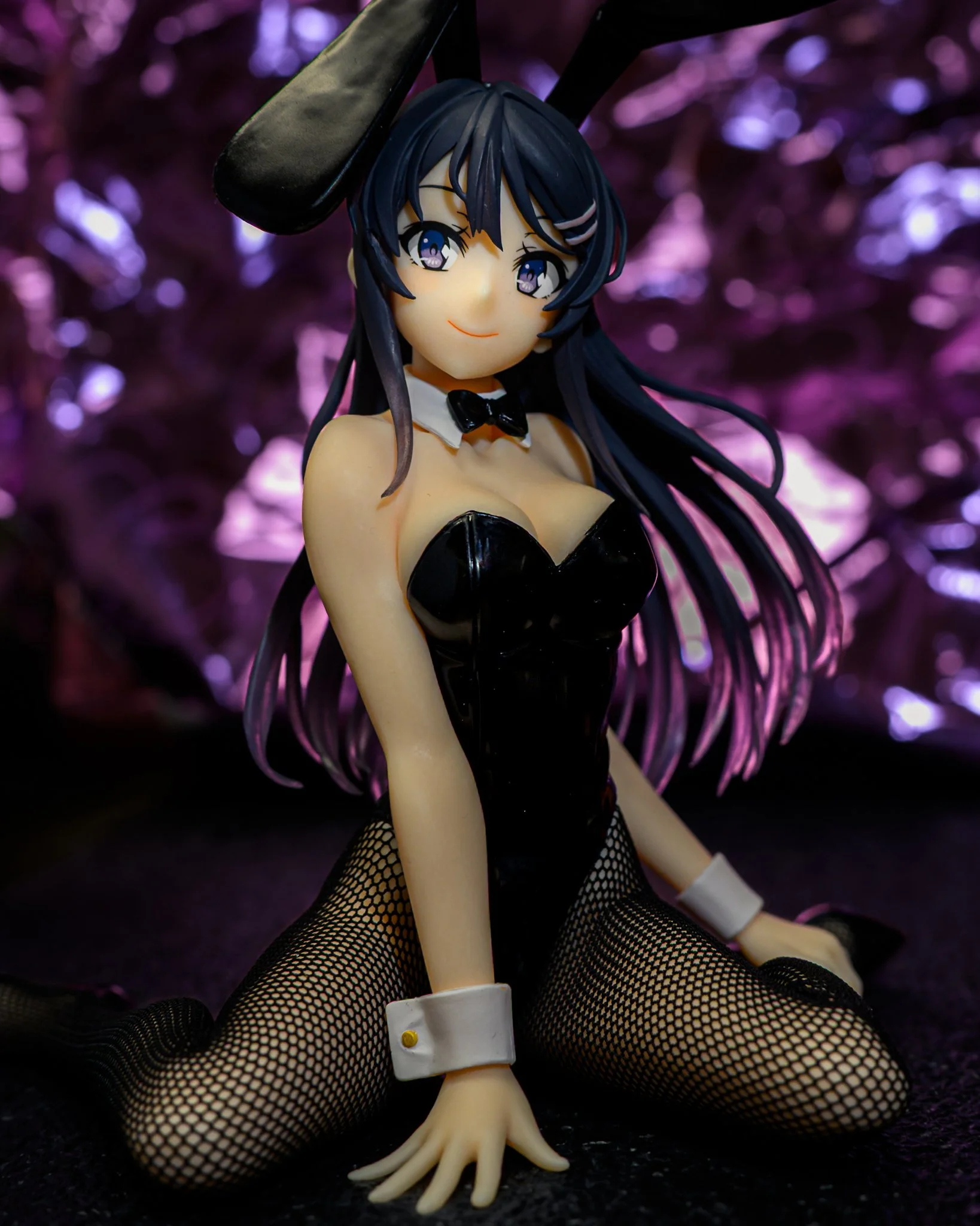 Anime-style figure of a young woman with long dark blue hair wearing a black bunny suit, fishnet stockings, and black rabbit ears. She sits posed on a dark surface with one hand forward, smiling softly. The background is a blurred purple bokeh scene 