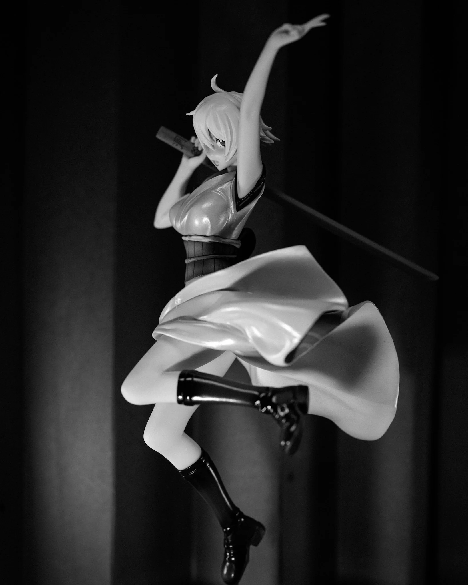 A black-and-white photograph of an anime-style figure posed dynamically with one arm raised overhead, holding a weapon behind her shoulder. Strong side lighting highlights the smooth contours of her arm and flowing skirt, while a dark background and 