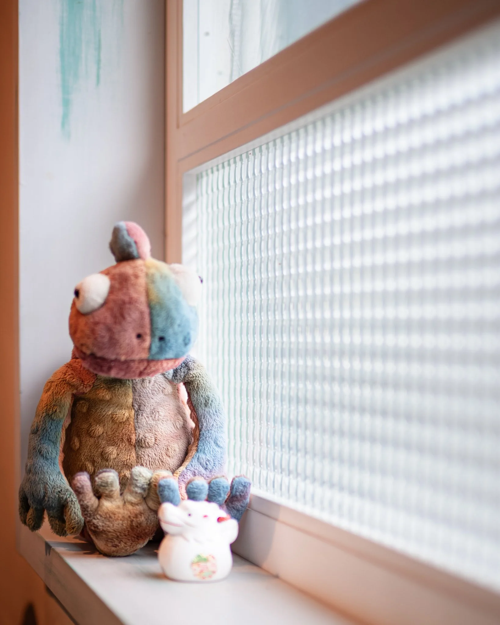 A colorful plush toy sits on a windowsill beside a small white ceramic figure, softly lit by daylight filtering through textured glass panels, creating a calm and intimate indoor scene.