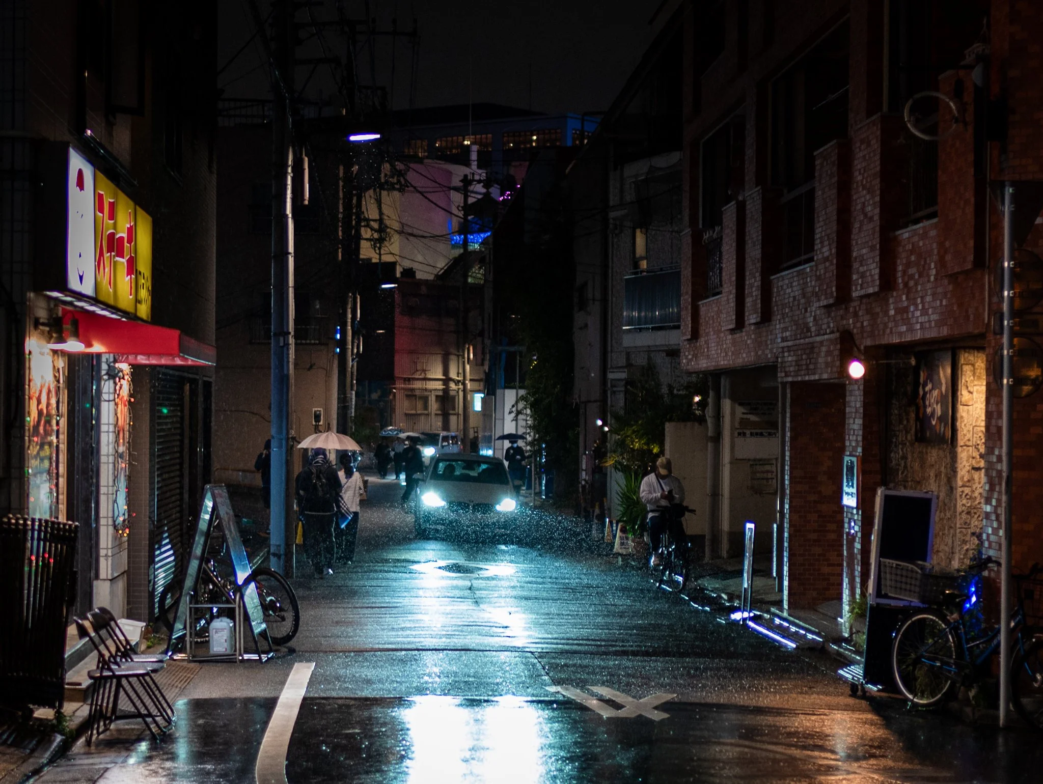 A rainy nighttime alley in a Japanese city, with wet pavement reflecting bright car headlights in the center. Pedestrians walk under umbrellas in the distance while a cyclist rides along the right side. Warm storefront lighting contrasts with cool st