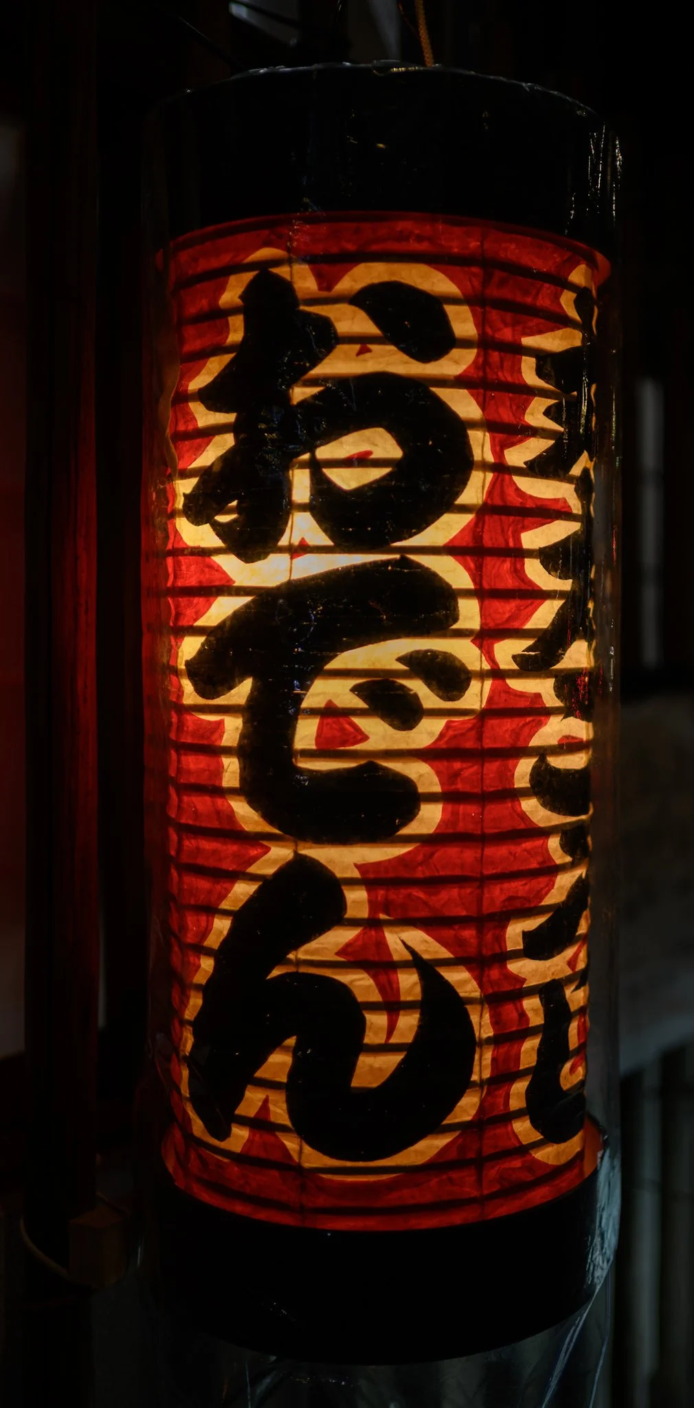 Close-up of a glowing red paper lantern at night, its warm amber light illuminating bold black Japanese calligraphy, with the surrounding background fading into darkness.