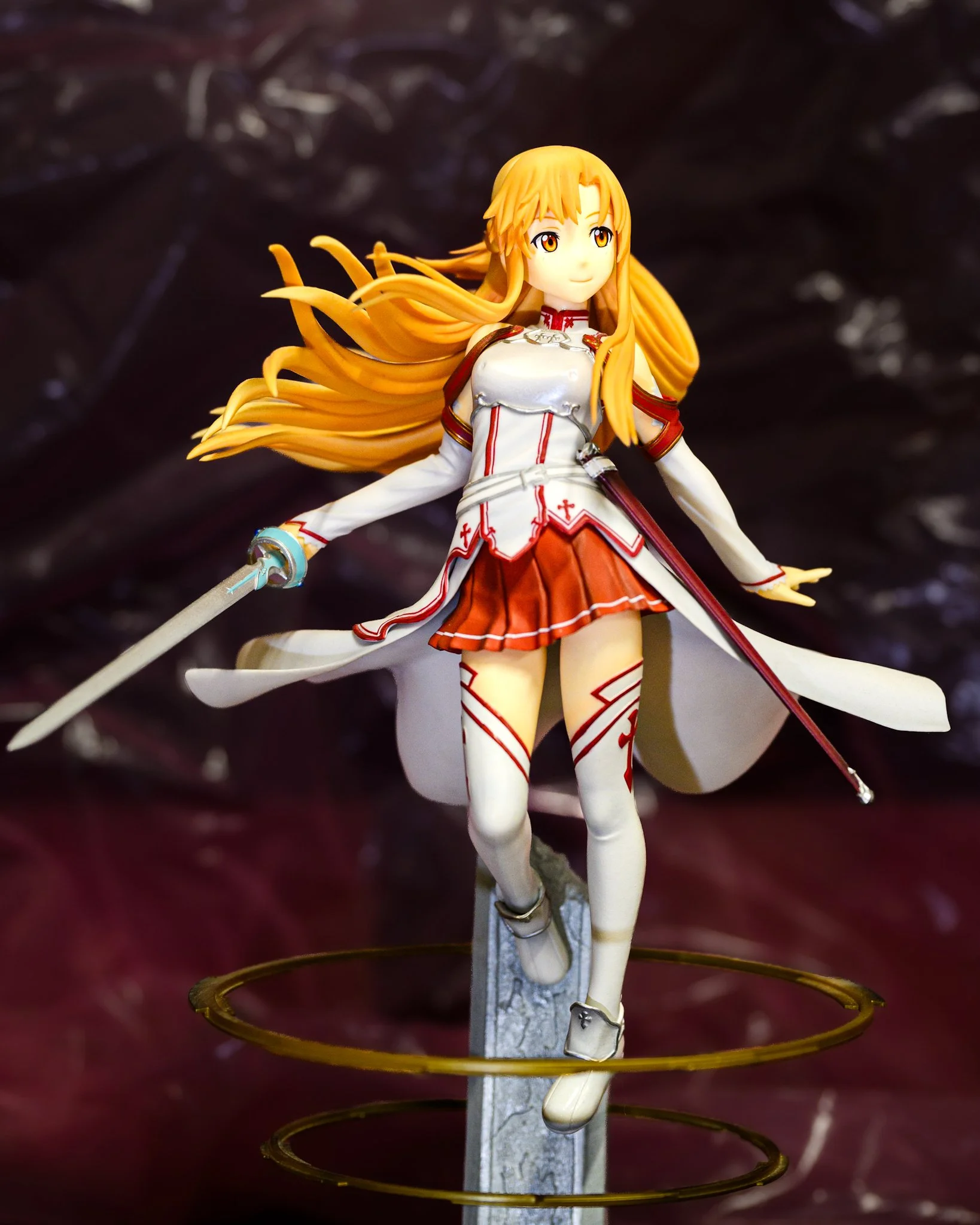 Anime figure of Asuna from Sword Art Online posed mid-stride with flowing golden hair and drawn sword, wearing her white-and-red Knights of the Blood uniform. She stands against a softly blurred burgundy background with subtle abstract textures, whil