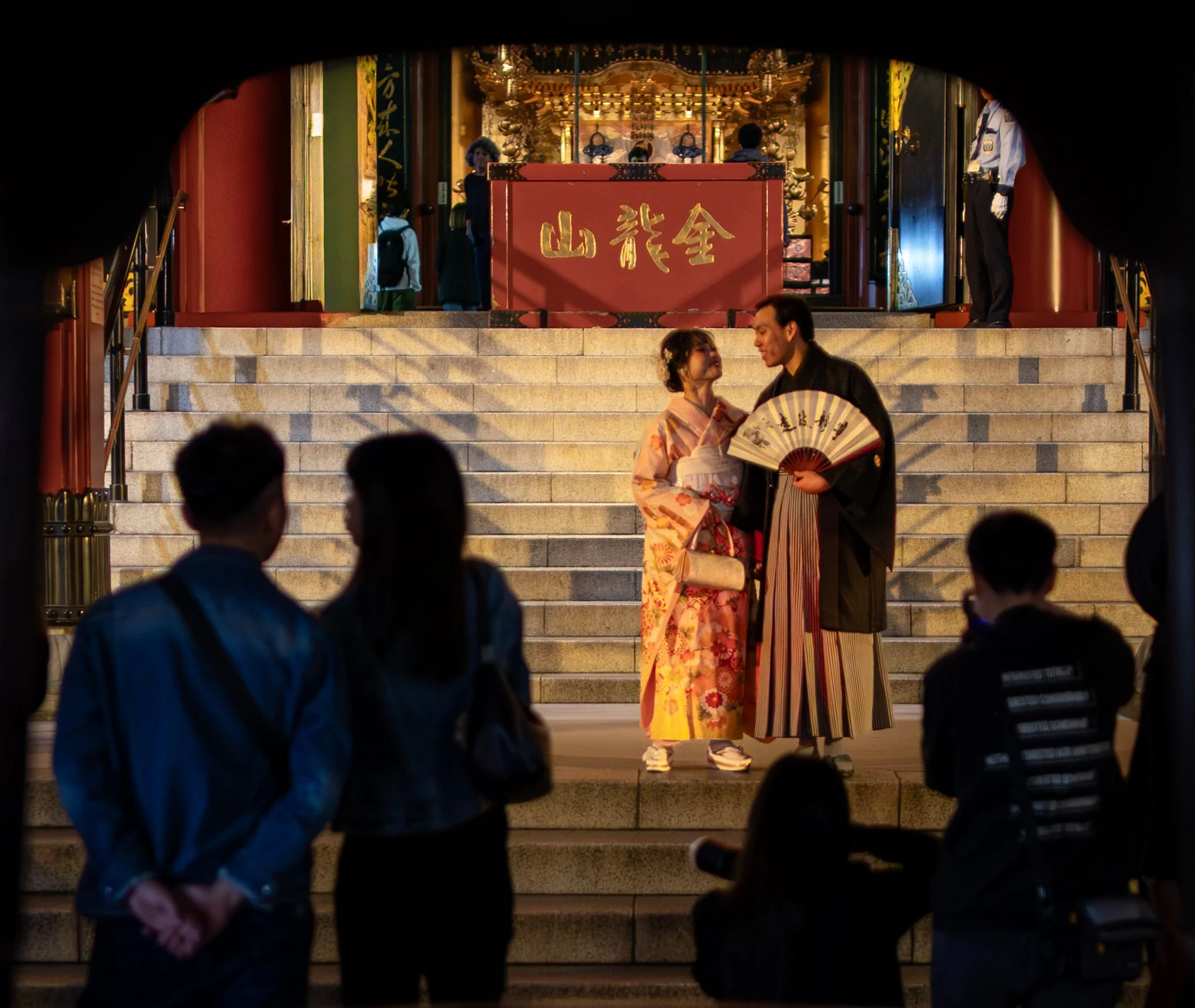 A warmly lit temple entrance at night frames a couple in traditional Japanese attire standing on stone steps. The woman wears a floral kimono and looks up at her companion, who wears a dark kimono and holds a folding fan. Behind them, a golden shrine