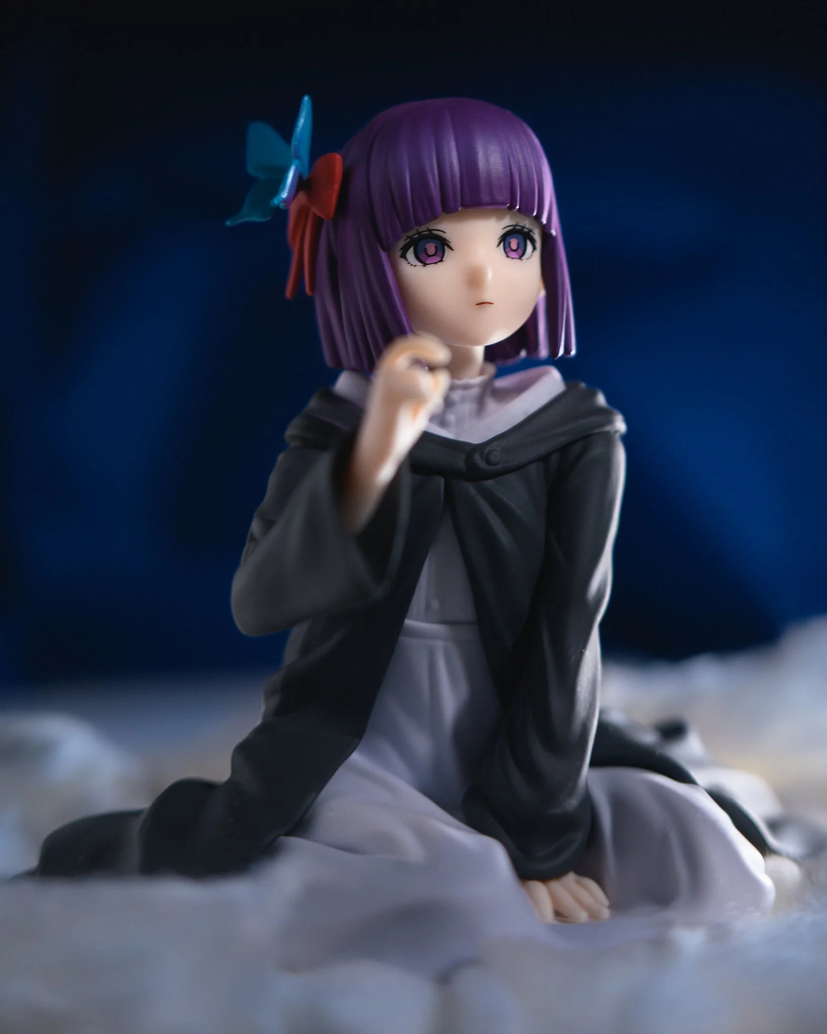 A detailed anime-style figure of a girl with short purple hair and a ribbon, seated on a soft, cloud-like surface. She wears a dark cloak over a white outfit and gazes slightly upward with a calm, contemplative expression against a deep blue backgrou