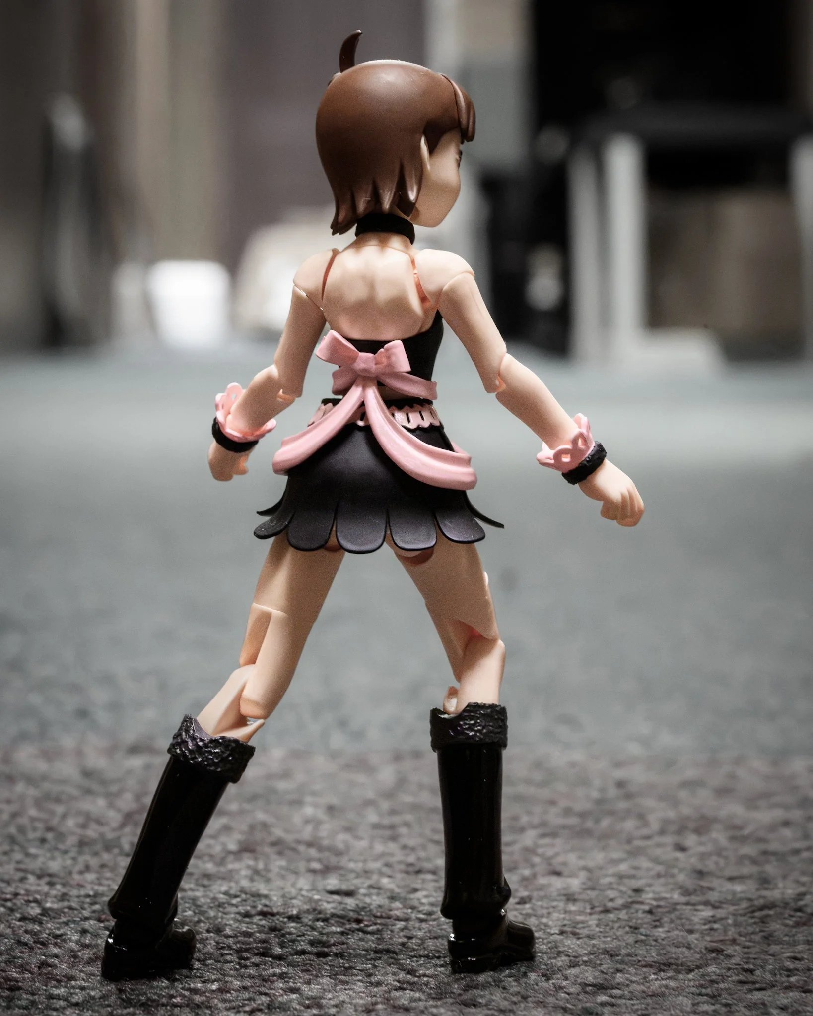 A muted-color photograph of an articulated anime figure standing with her back to the camera on a carpeted floor. She faces a dark hallway and a softly illuminated doorway, while the subdued colors and gentle lighting emphasize her pink ribbon and cr