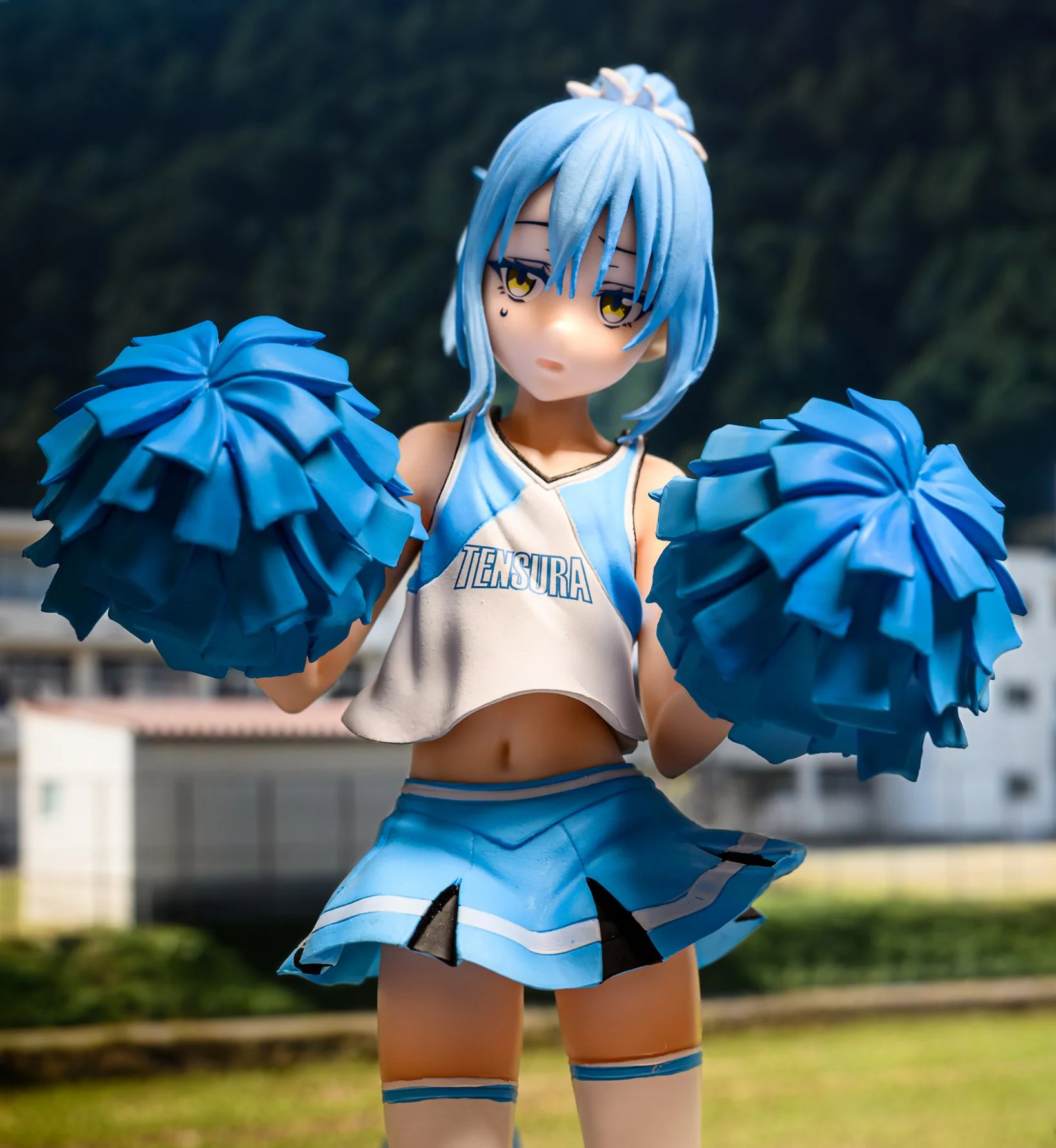 A blue-haired anime-style figure in a cheerleader outfit stands outdoors holding two large blue pom-poms. The figure wears a white crop top with “TENSURA” printed on it and a blue skirt, with a blurred background of greenery and buildings behind.