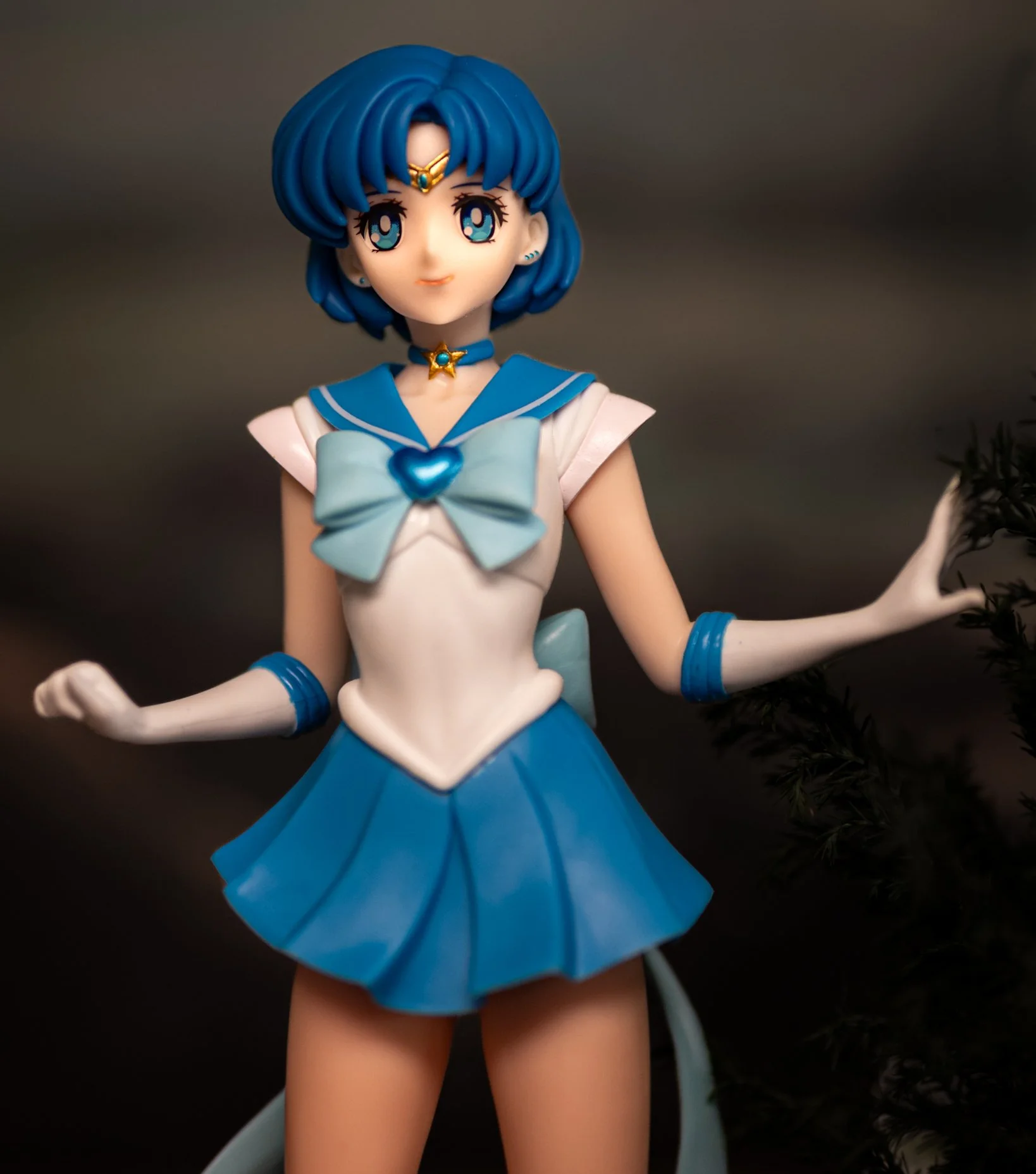 Anime-style figure of Sailor Mercury with short blue hair and teal eyes, wearing a blue-and-white sailor uniform with a large bow and star choker, posed with arms slightly out against a softly blurred neutral background with a hint of greenery on the