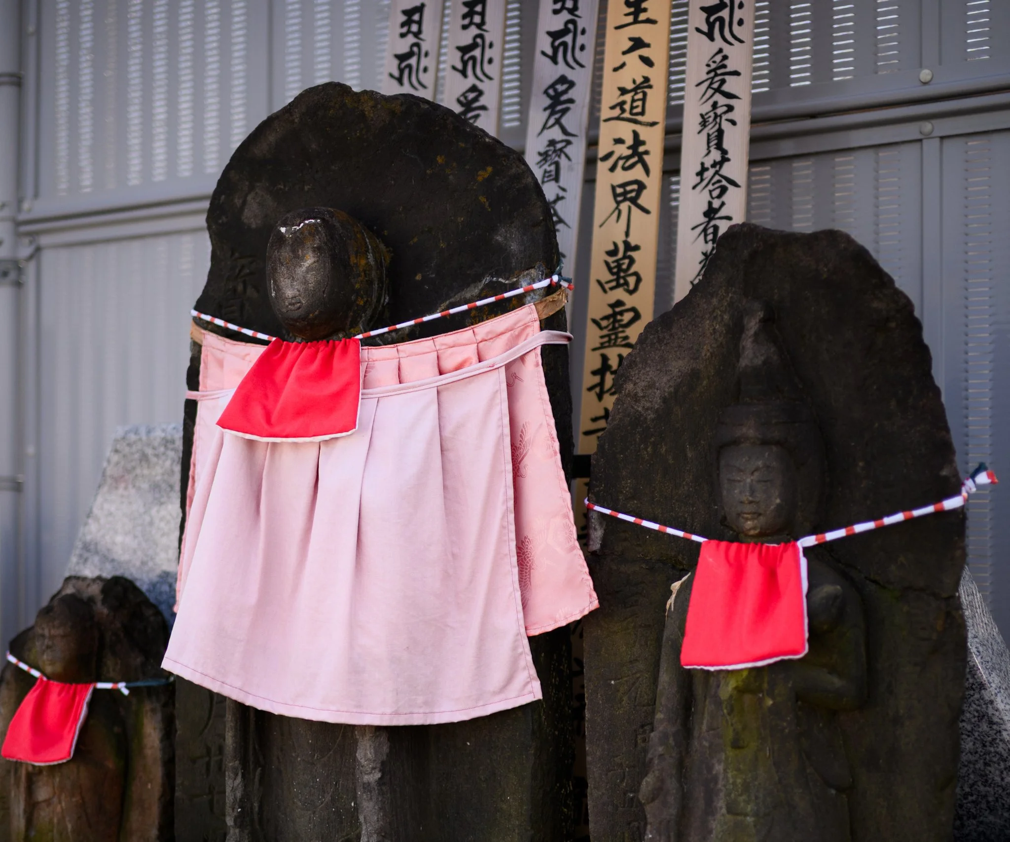 Close-up of weathered stone Jizō statues in Tokyo, each dressed in red bibs and one wearing a pale pink cloth garment. The figures stand side by side, while wooden prayer tablets with Japanese calligraphy rise behind them.