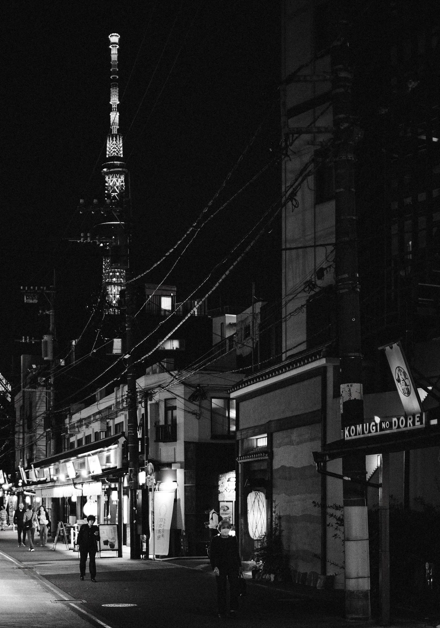 A black-and-white nighttime street scene in a Japanese neighborhood, with pedestrians walking along a softly lit road lined with small shops and buildings. Overhead power lines lead toward a brightly illuminated tower in the distance, while deep shad