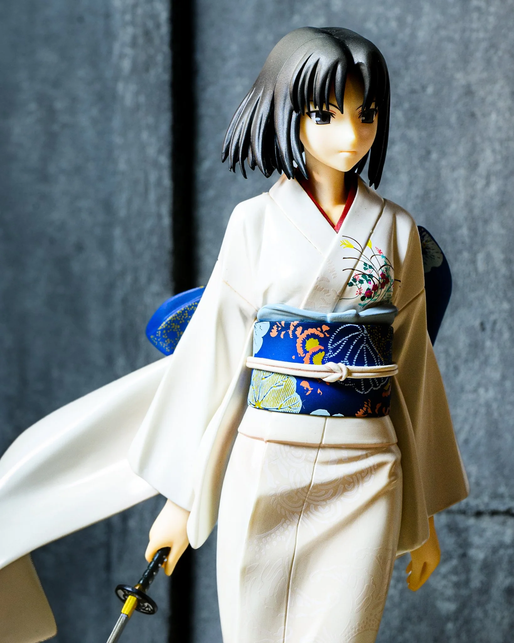 Shiki Ryougi anime figure in a white floral kimono holding a katana, standing before a weathered concrete wall with dramatic side lighting and a sweeping sleeve creating a cinematic composition.