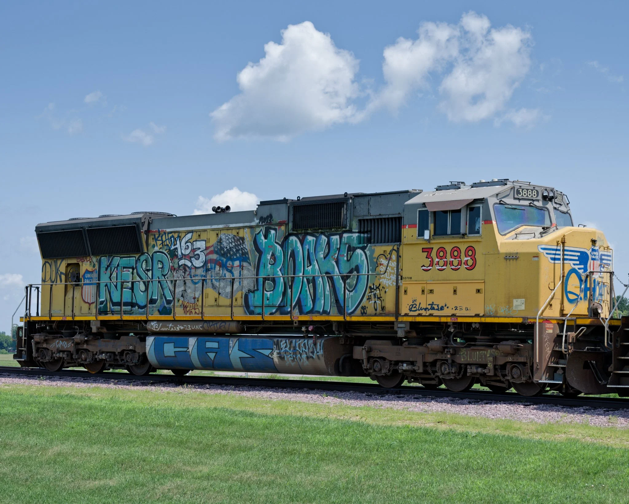 Broadside view of Union Pacific locomotive 3888 beneath a partly cloudy blue sky, its weathered yellow body extensively covered with colorful graffiti in turquoise, blue, purple, green, white, and black. Graffiti also covers the fuel tank and lower e