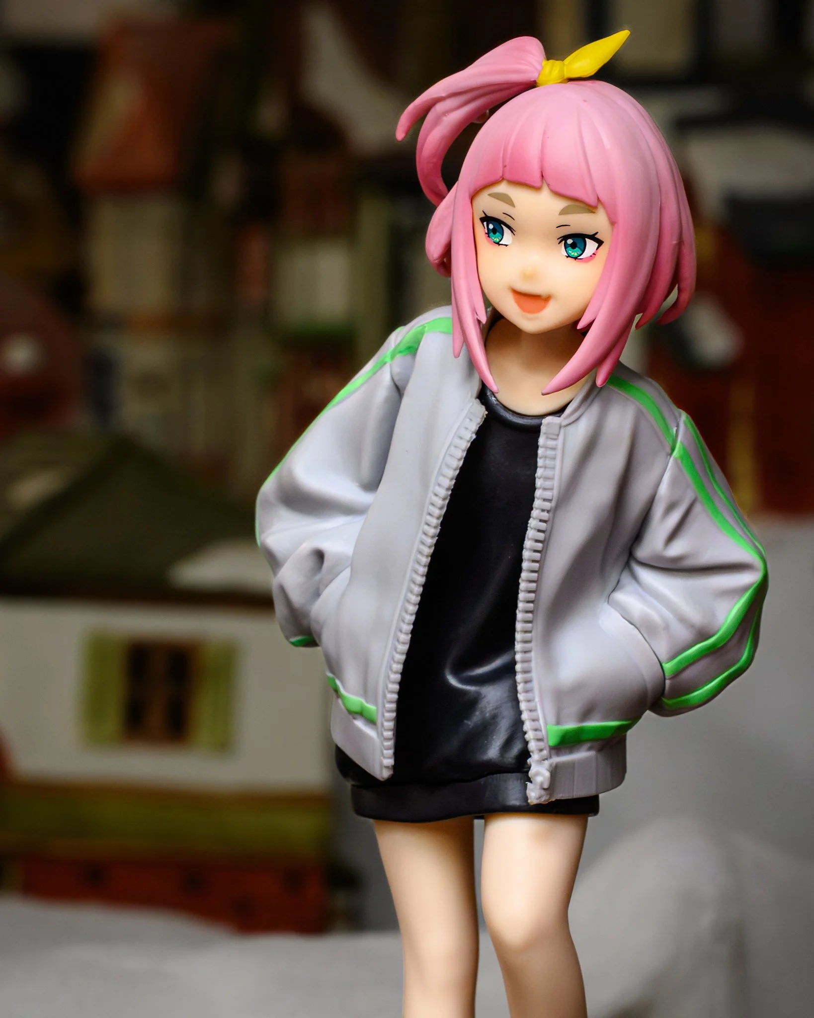 A stylized anime figure with short pink hair and a yellow hair accessory stands in front of a softly blurred miniature Christmas village scene. The shallow depth of field and lighting create the illusion that the figure is life-sized within a distant