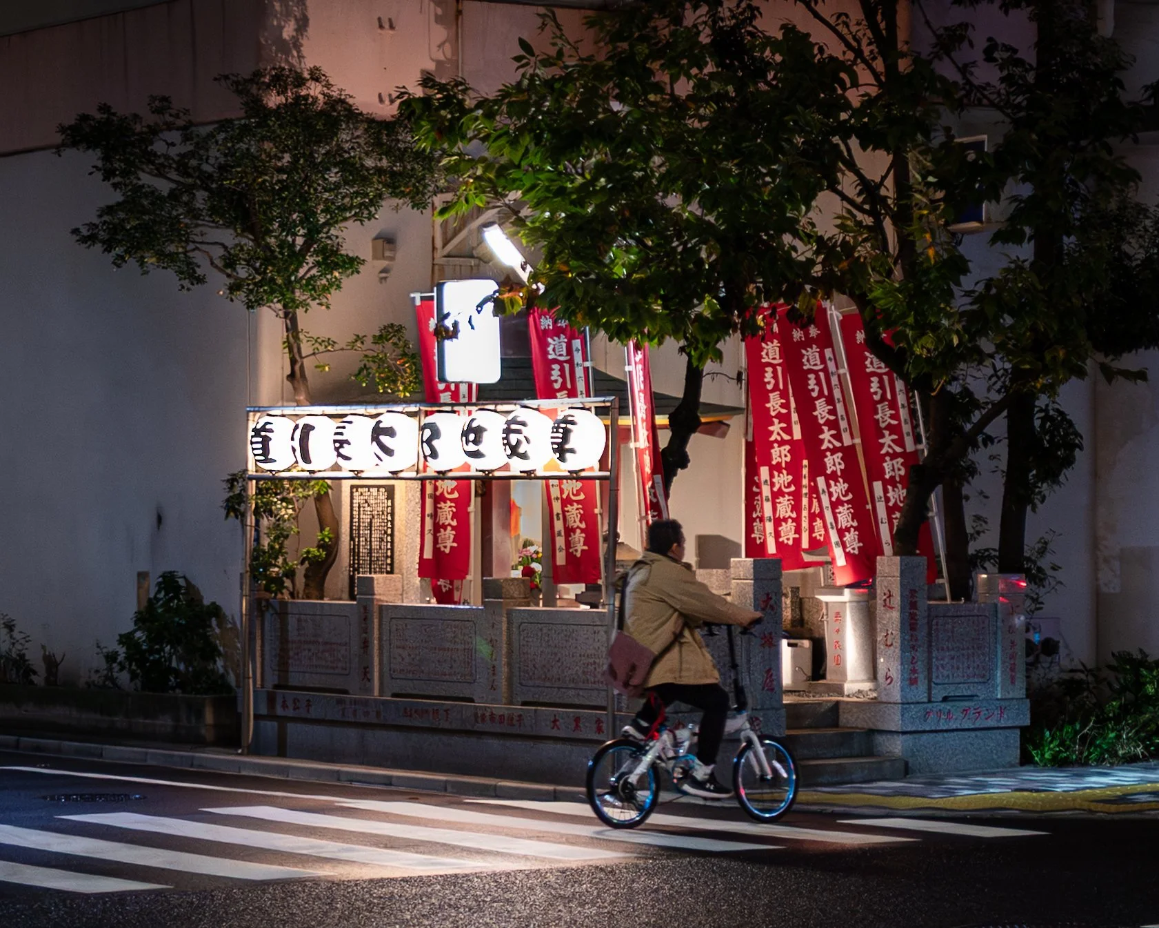 At night, a cyclist rides across a crosswalk past a small illuminated roadside shrine adorned with red banners and paper lanterns, partially framed by trees against a softly lit building.