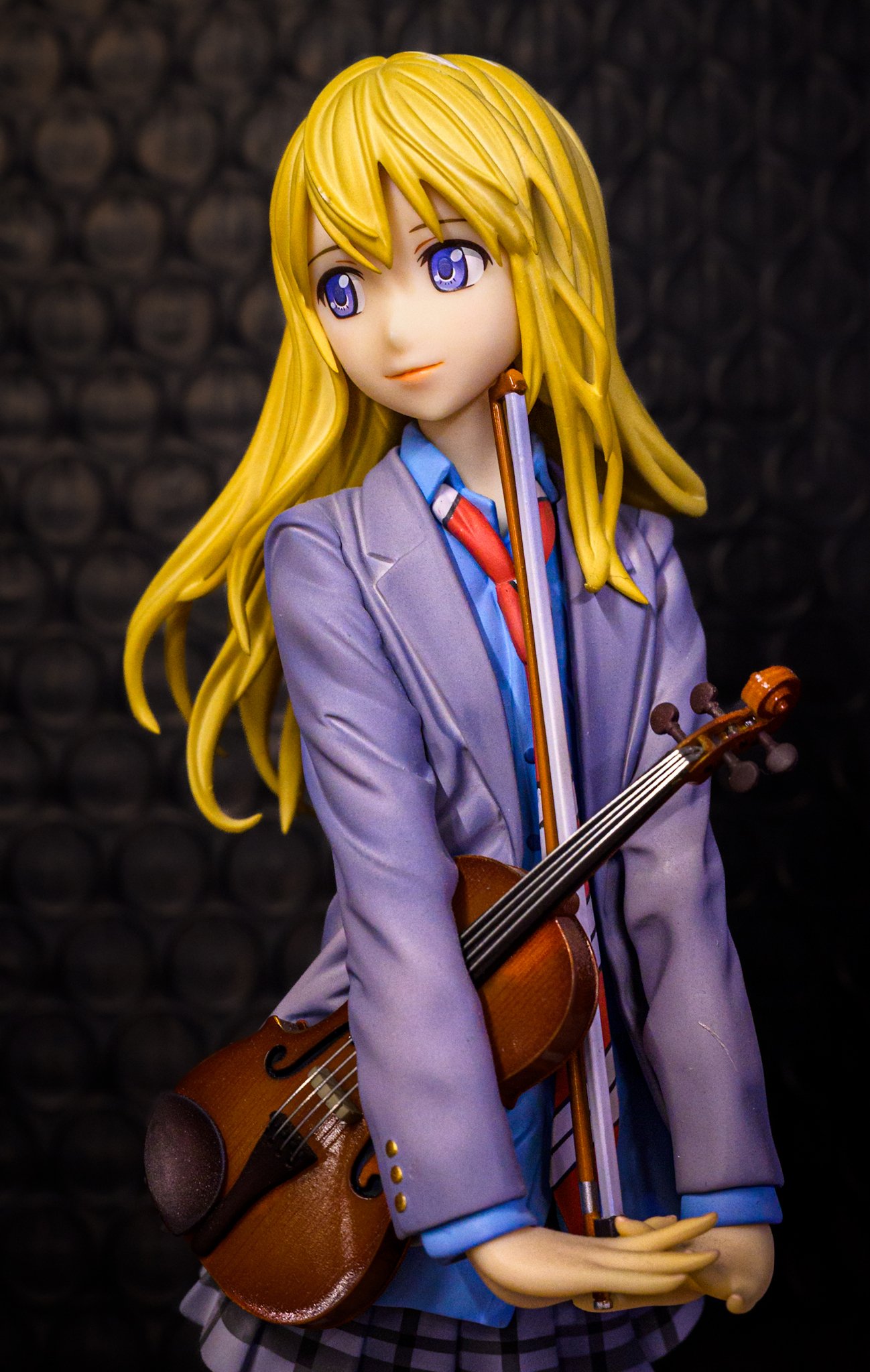 Anime-style figure of a blonde girl in a school uniform holding a violin and bow, posed against a dark patterned background with soft, directional lighting.
