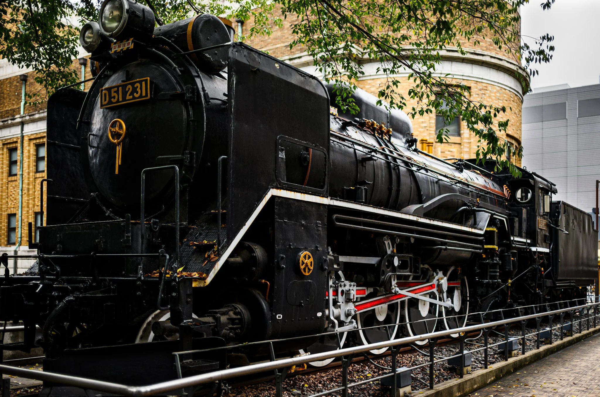 Large black steam locomotive displayed outdoors in Ueno beside a museum building, photographed from a low angle under green tree branches. White wheels and red connecting rods contrast with the dark engine body, while curved brick architecture rises 