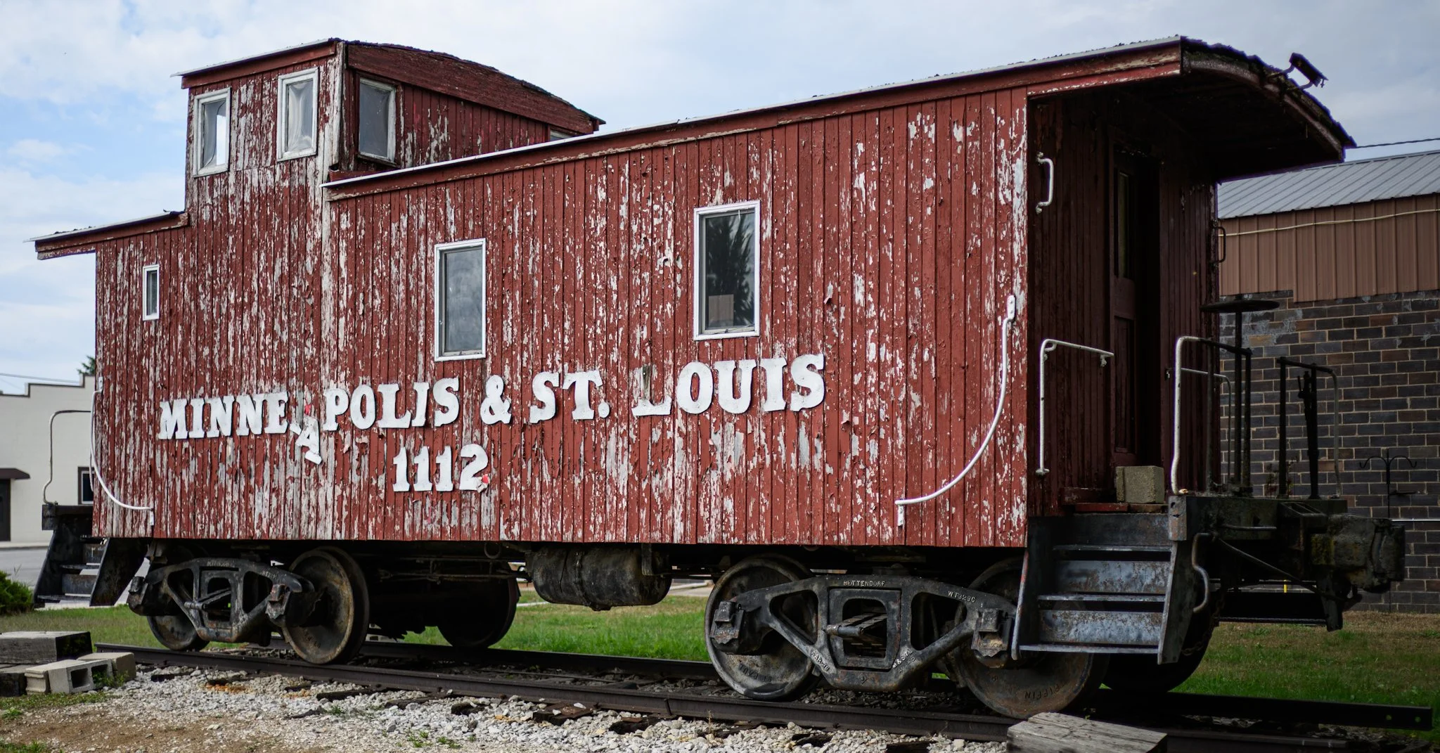 A weathered red Minneapolis & St. Louis caboose numbered 1112 sits on short display tracks beside a modern building. Peeling red paint and large white lettering highlight the preserved railcar’s railroad history, while dark wheels, worn wood siding, 