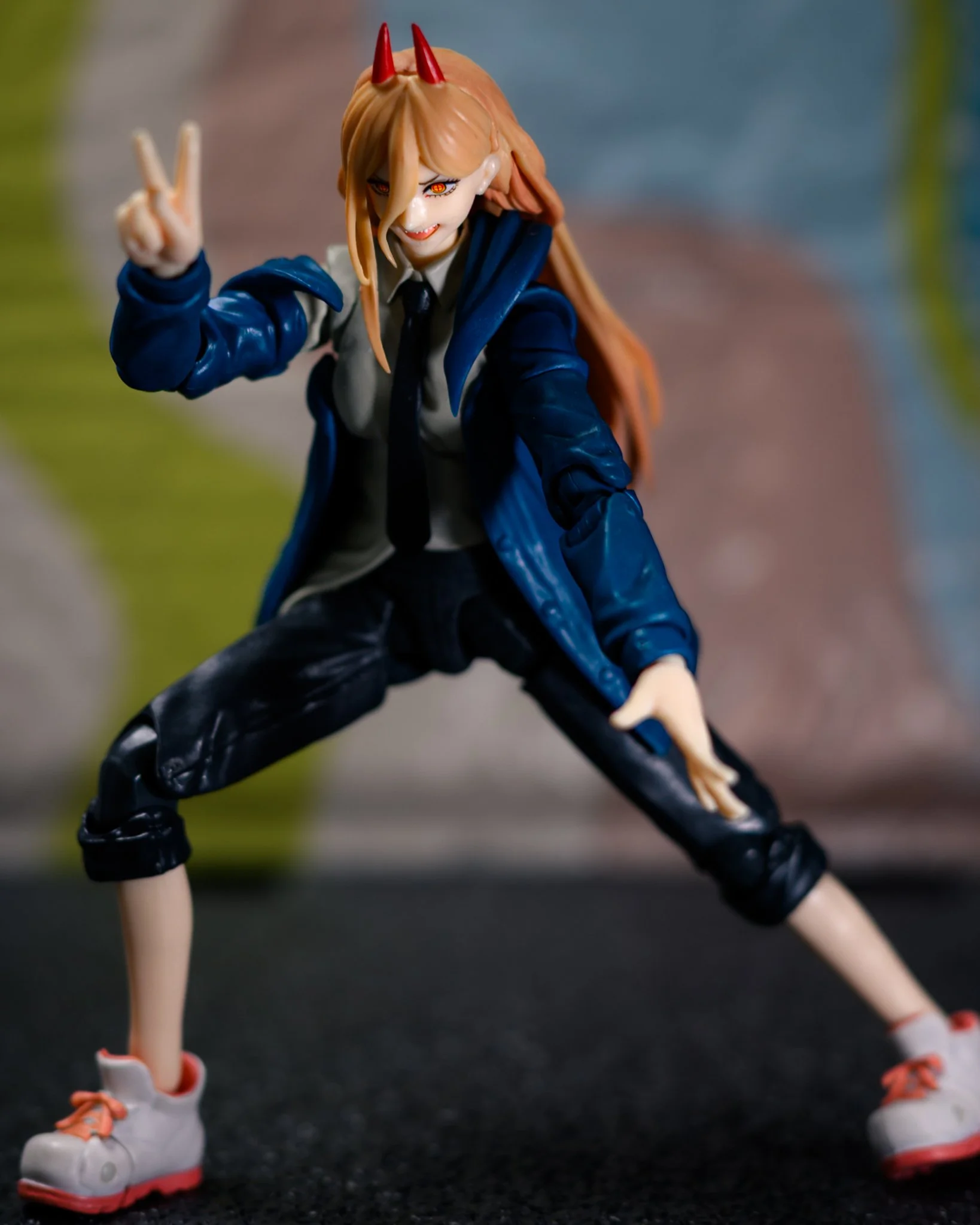 Close-up photo of a Power action figure from Chainsaw Man posed in a wide crouched stance on a dark textured surface. She wears a blue jacket, white shirt, black tie, cropped black pants, and white sneakers with red accents. One hand flashes a peace 