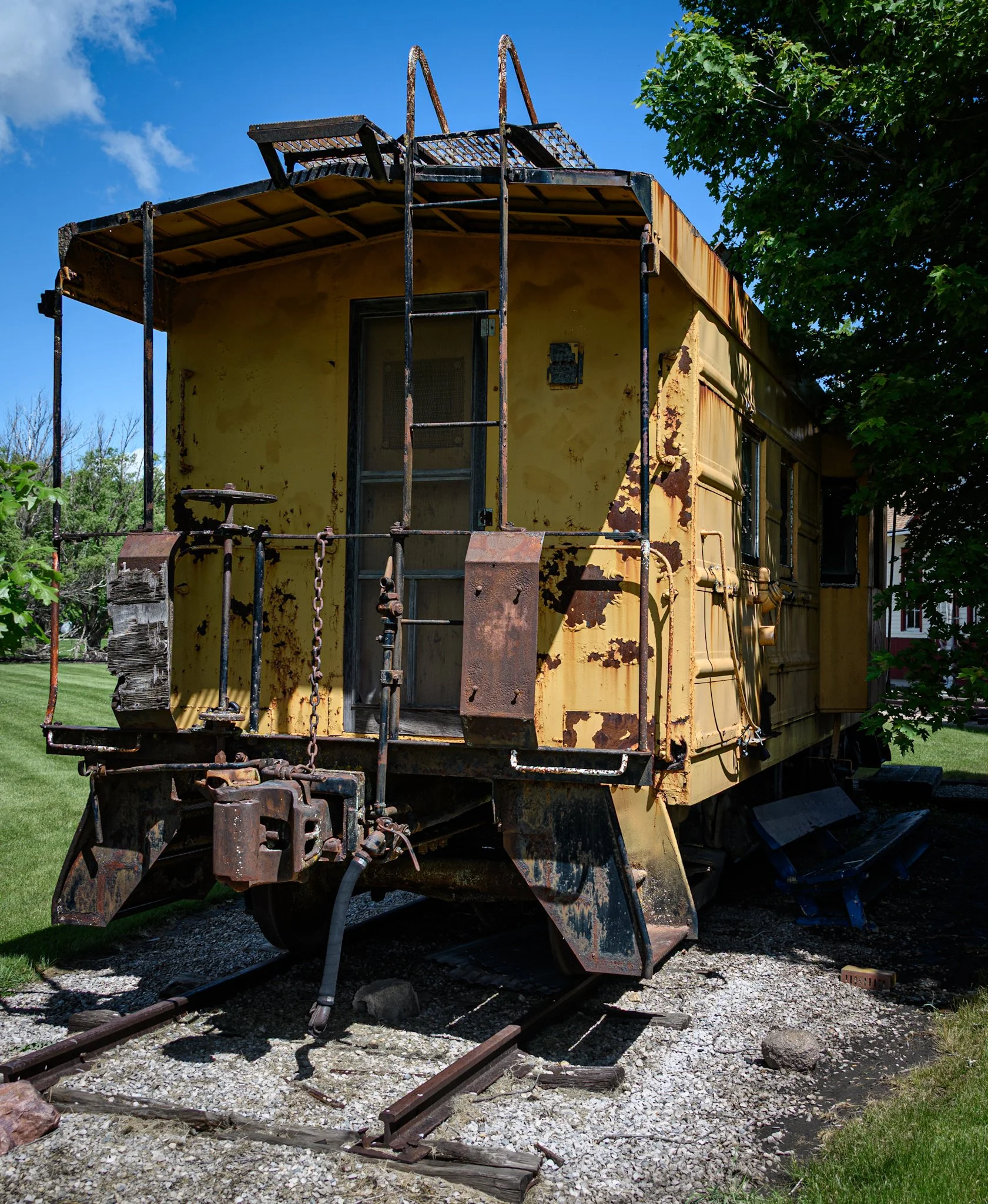 A weathered yellow Milwaukee Road caboose sits on short display tracks beneath a vivid blue summer sky. Rust covers the coupler, ladder, roof platform, and rear platform, while deep tree shadows frame one side and emphasize the age and texture of the