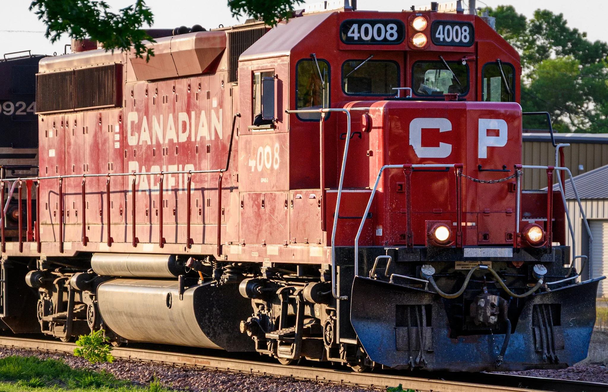 A bright red Canadian Pacific locomotive numbered 4008 approaches along the tracks in warm evening light. Green grass fills the foreground, while industrial buildings and leafy trees frame the background, giving the train a strong urban rail yard pre