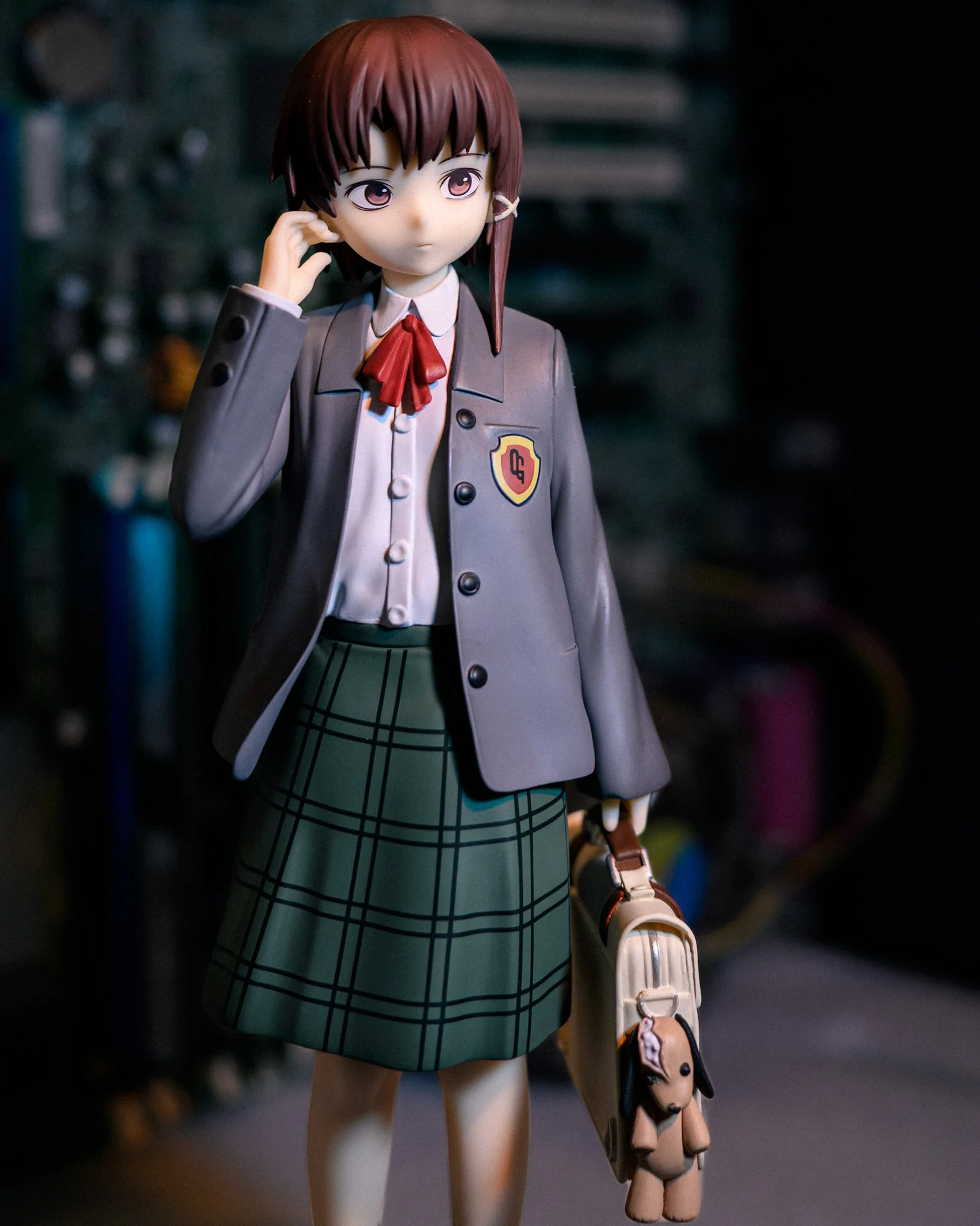 A schoolgirl figure of Lain Iwakura stands against a softly blurred computer motherboard backdrop, lit by moody blue-green shadows and warm highlights. She wears a gray school blazer with a red ribbon tie and green plaid skirt, holding a beige school