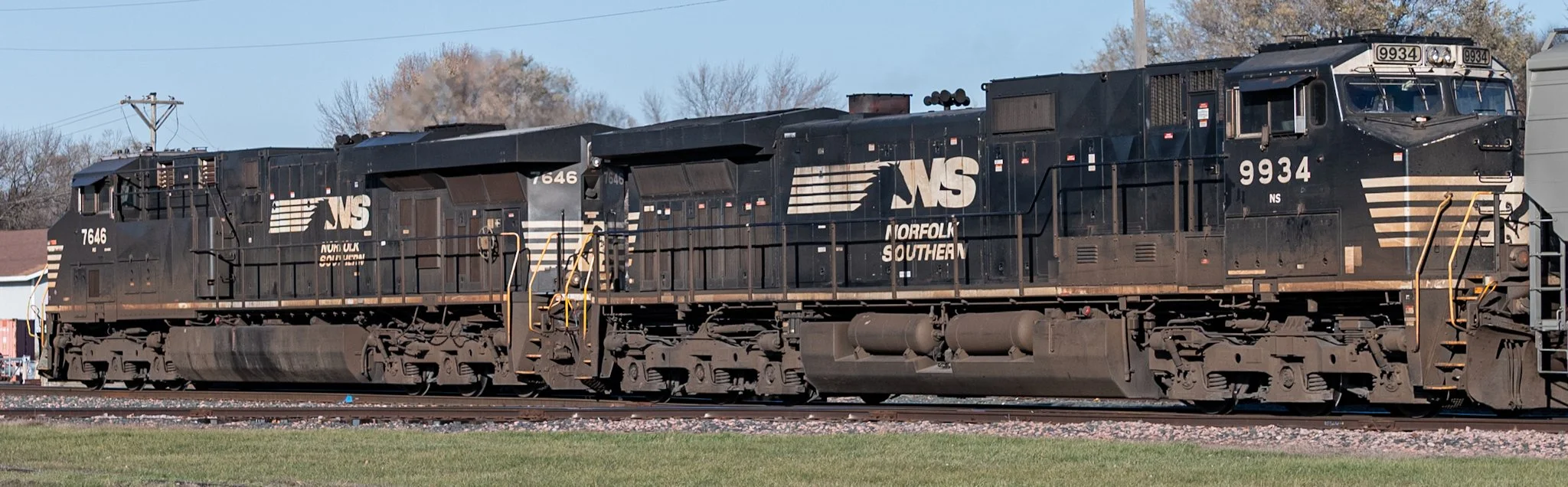 Two black Norfolk Southern locomotives move along the tracks in side profile under a clear blue sky. Their white NS logos stand out against the dark engines, while grass and gravel in the foreground emphasize the long, heavy industrial presence of th